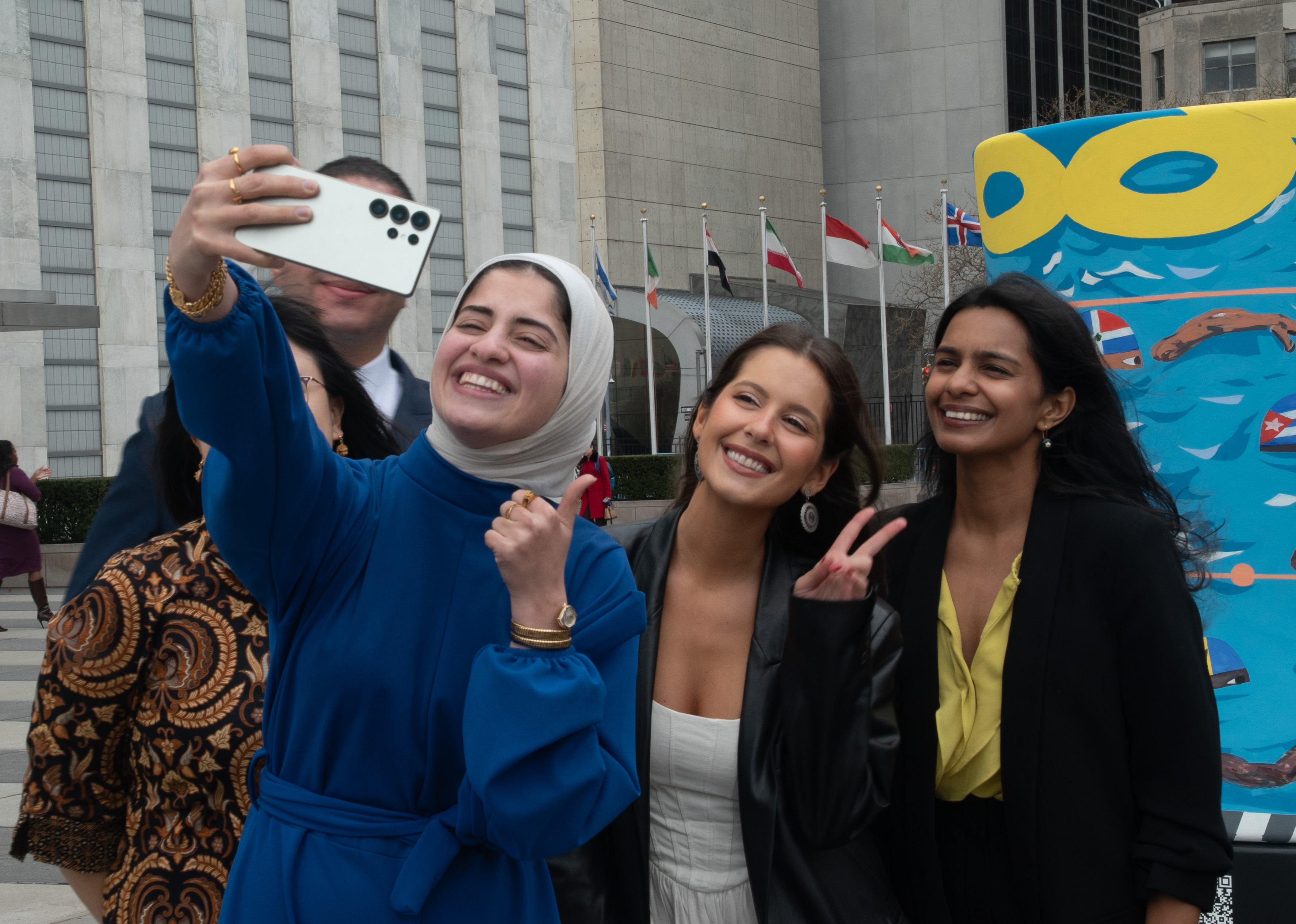 Four people posing in front of a city building; blue-clad person holds a white card.