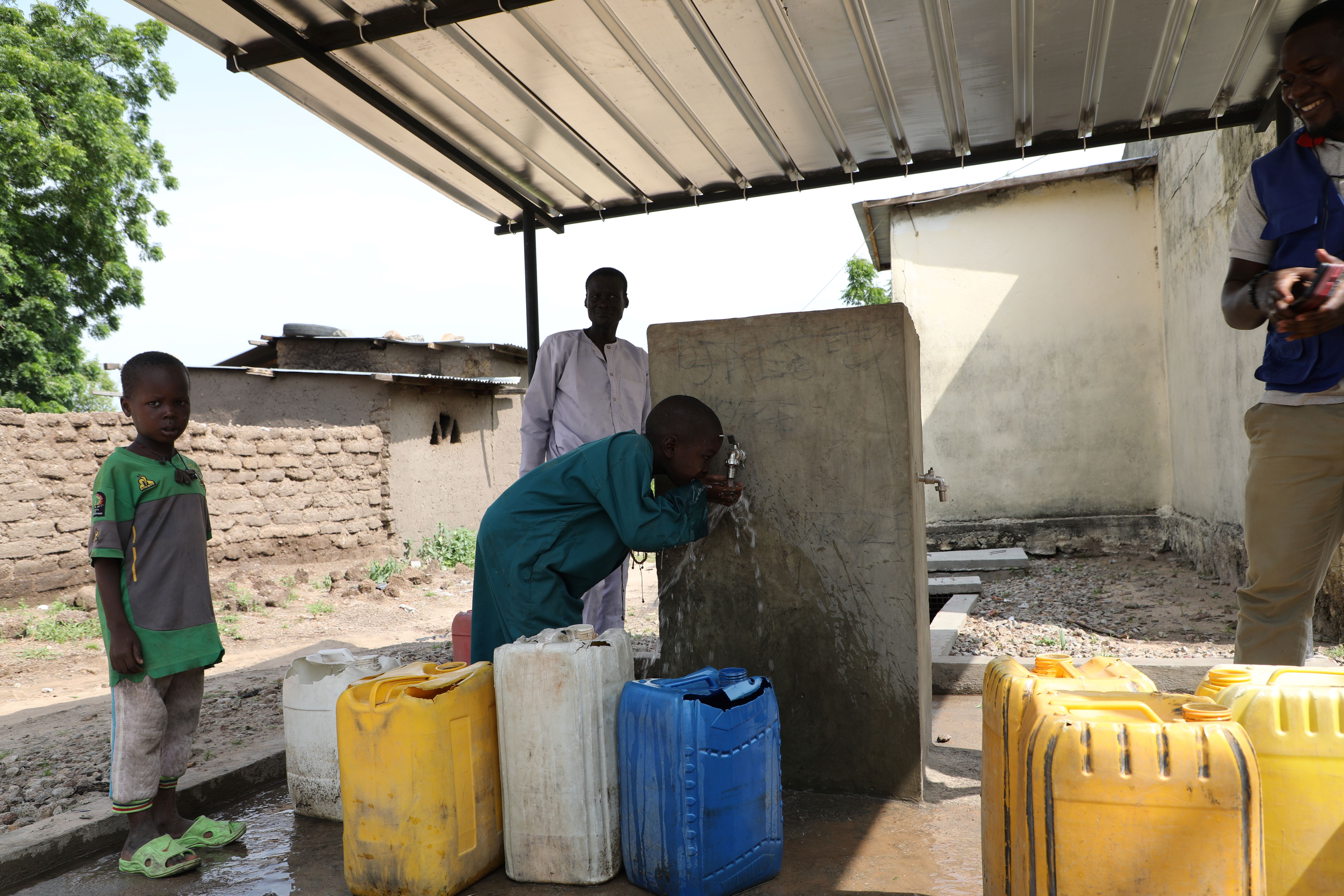 Man fills yellow and blue jerry cans with water from a hand pump under a shade; a child watches.
