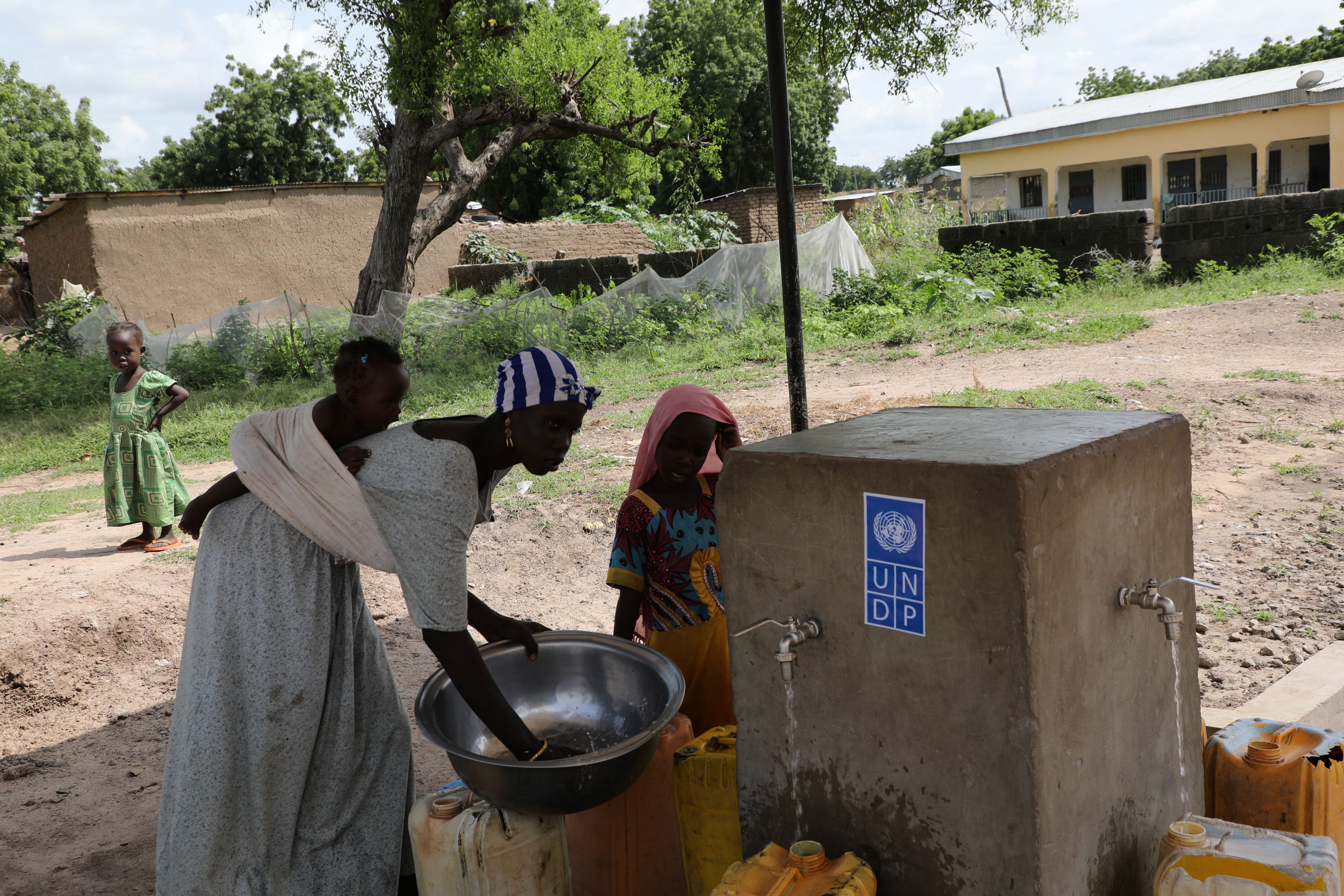 People collect water from a hand pump beside a concrete cistern in a rural yard.