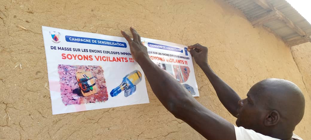 Man holds two posters with French 'Soyons vigilants' slogans and color photos on a clay wall.