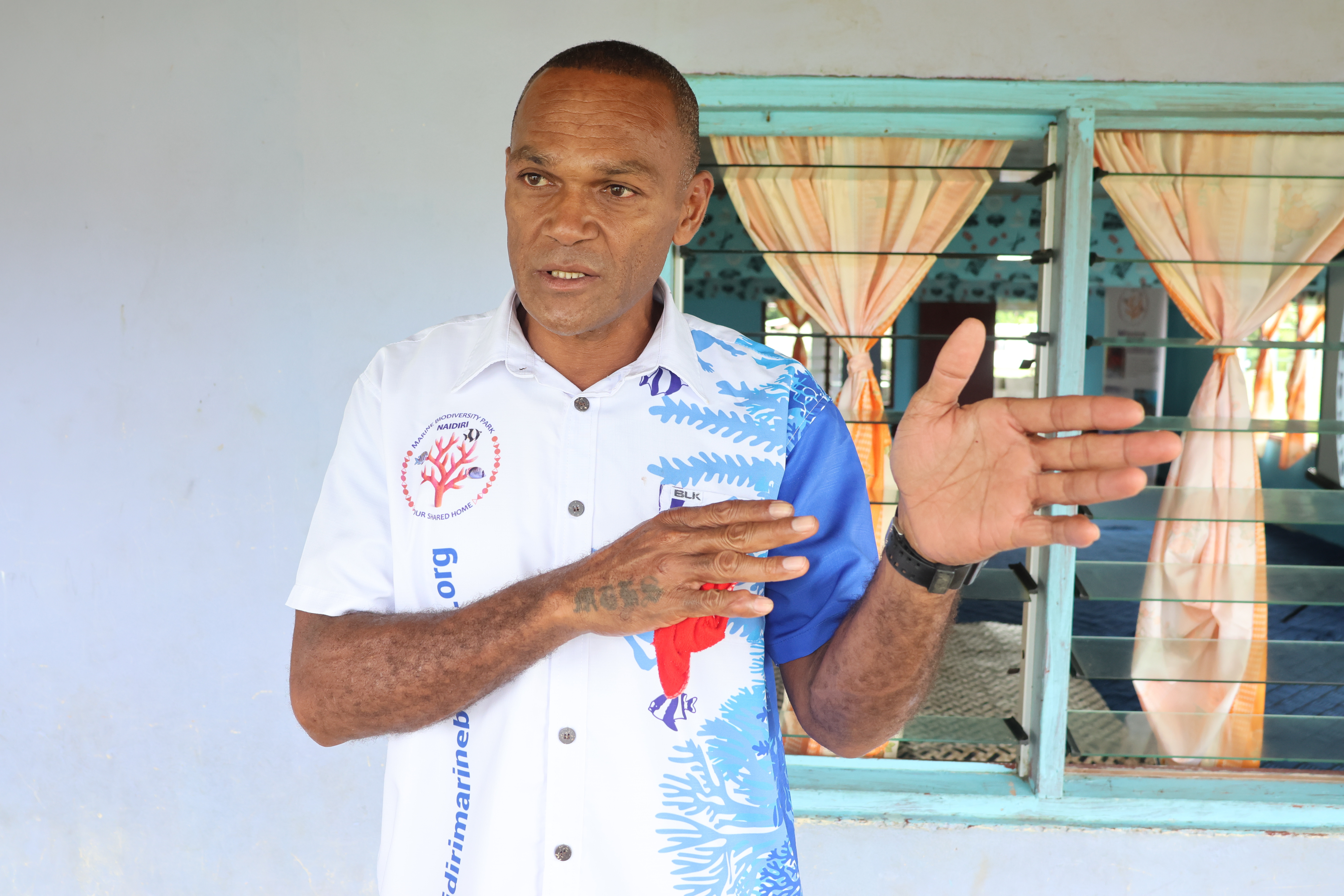 Man in a white polo shirt with blue accents stands near a window, gesturing with an open hand.