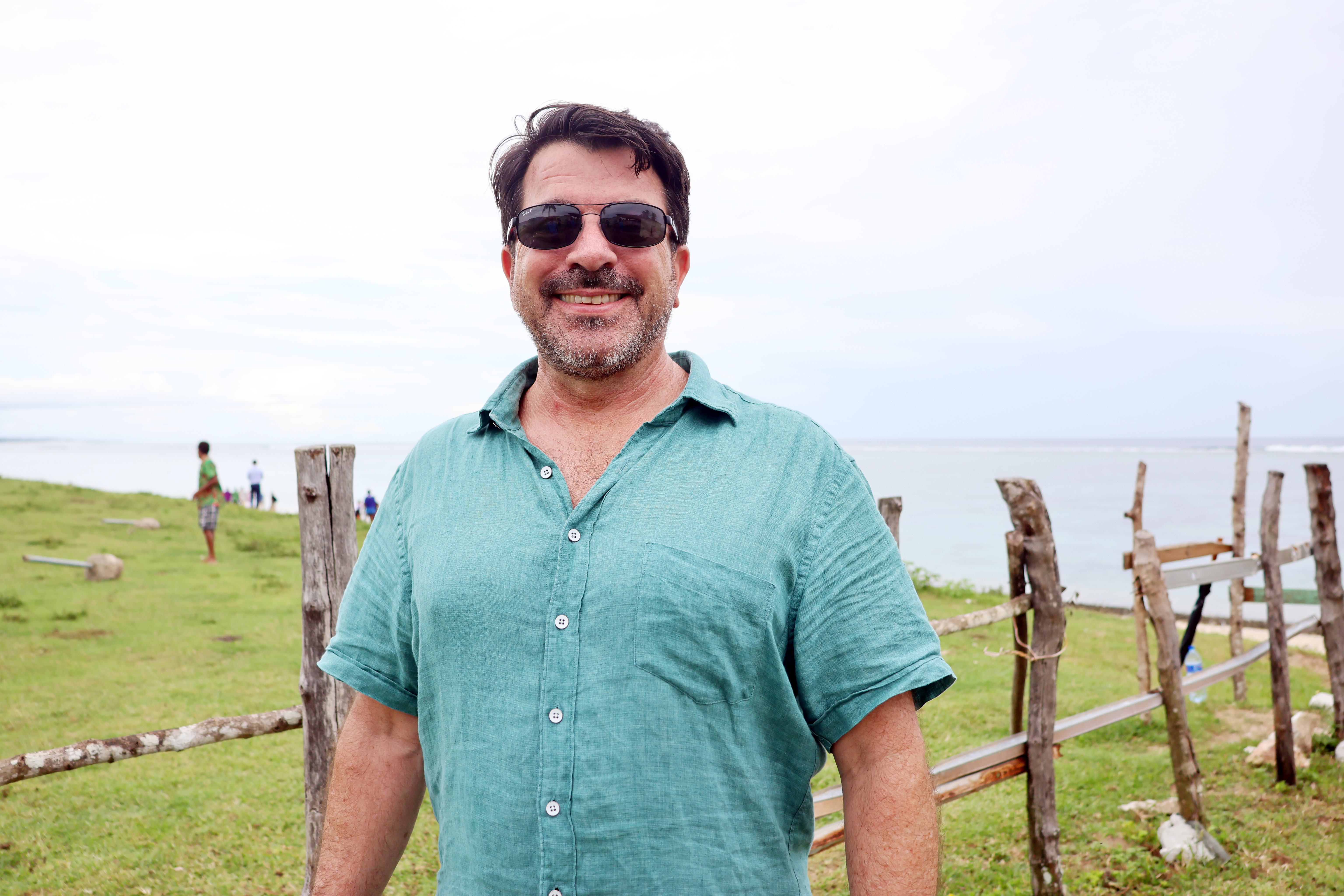 Man in teal shirt stands in a grassy field beside a wooden fence.