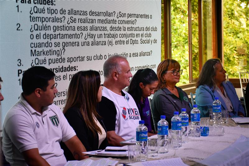 Photo: five panelists at a table with water bottles, outdoors, and a projected agenda.