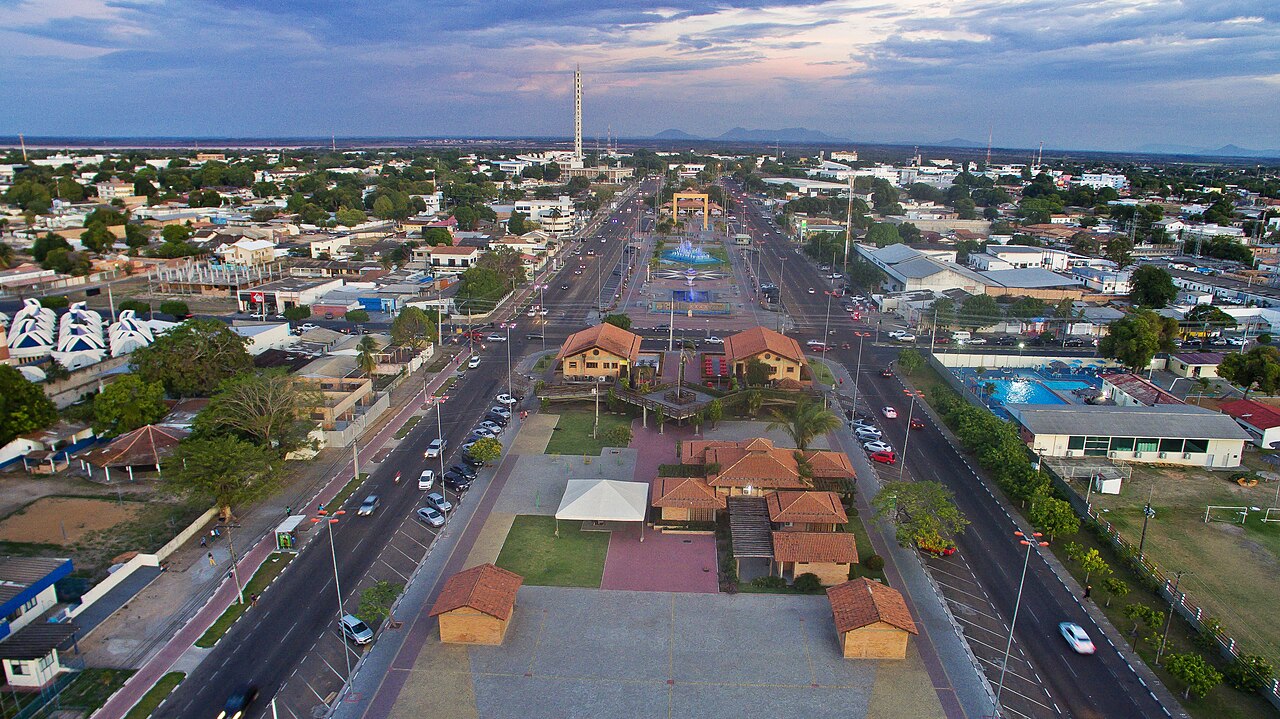 An aerial view of a wide suburban avenue with houses on both sides, palm trees, and a blue sky.