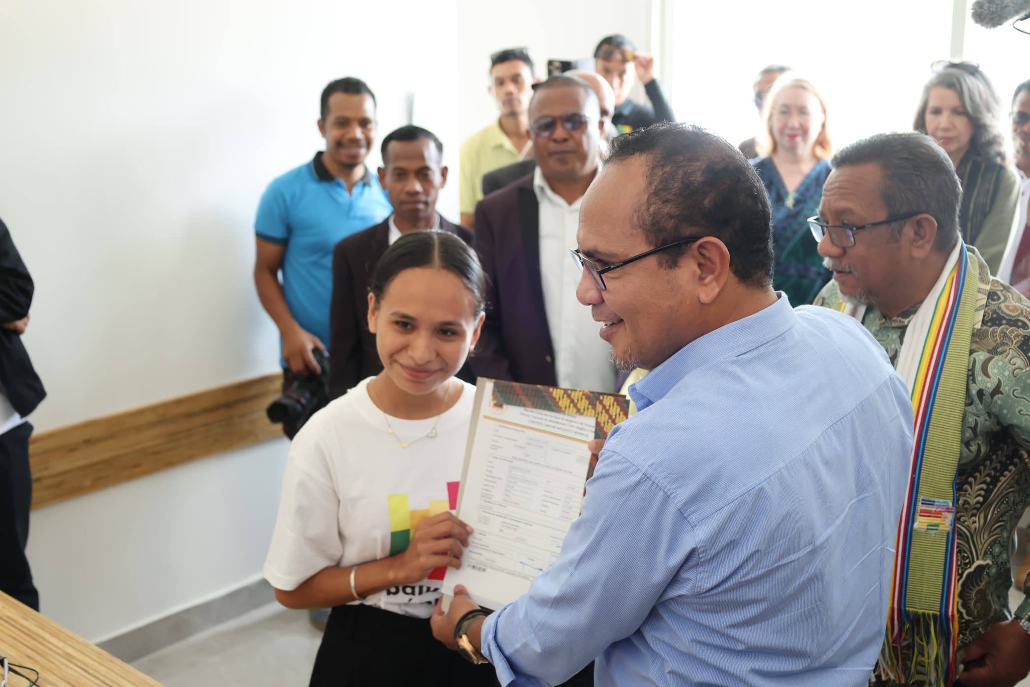 Man in a blue shirt presents a certificate to a recipient at a ceremony, surrounded by onlookers.