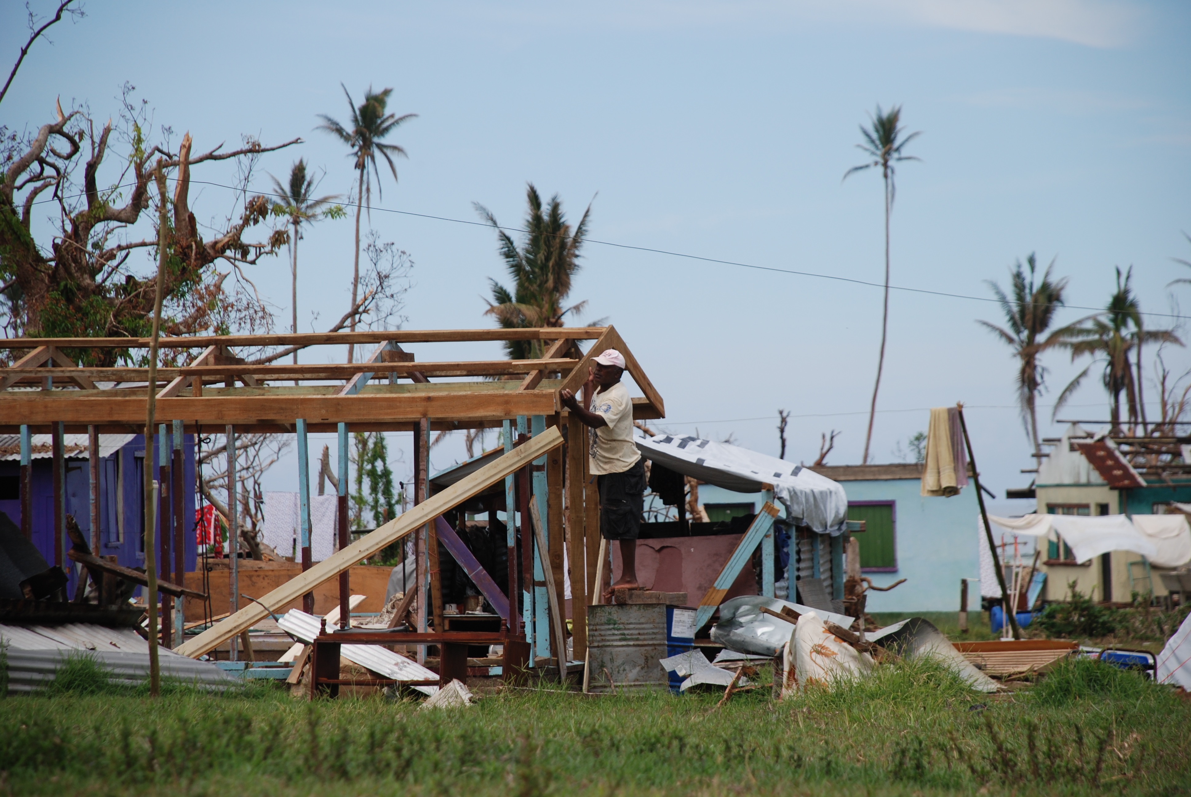 Damaged makeshift settlement with collapsed frames, tarps, and debris near palm trees.