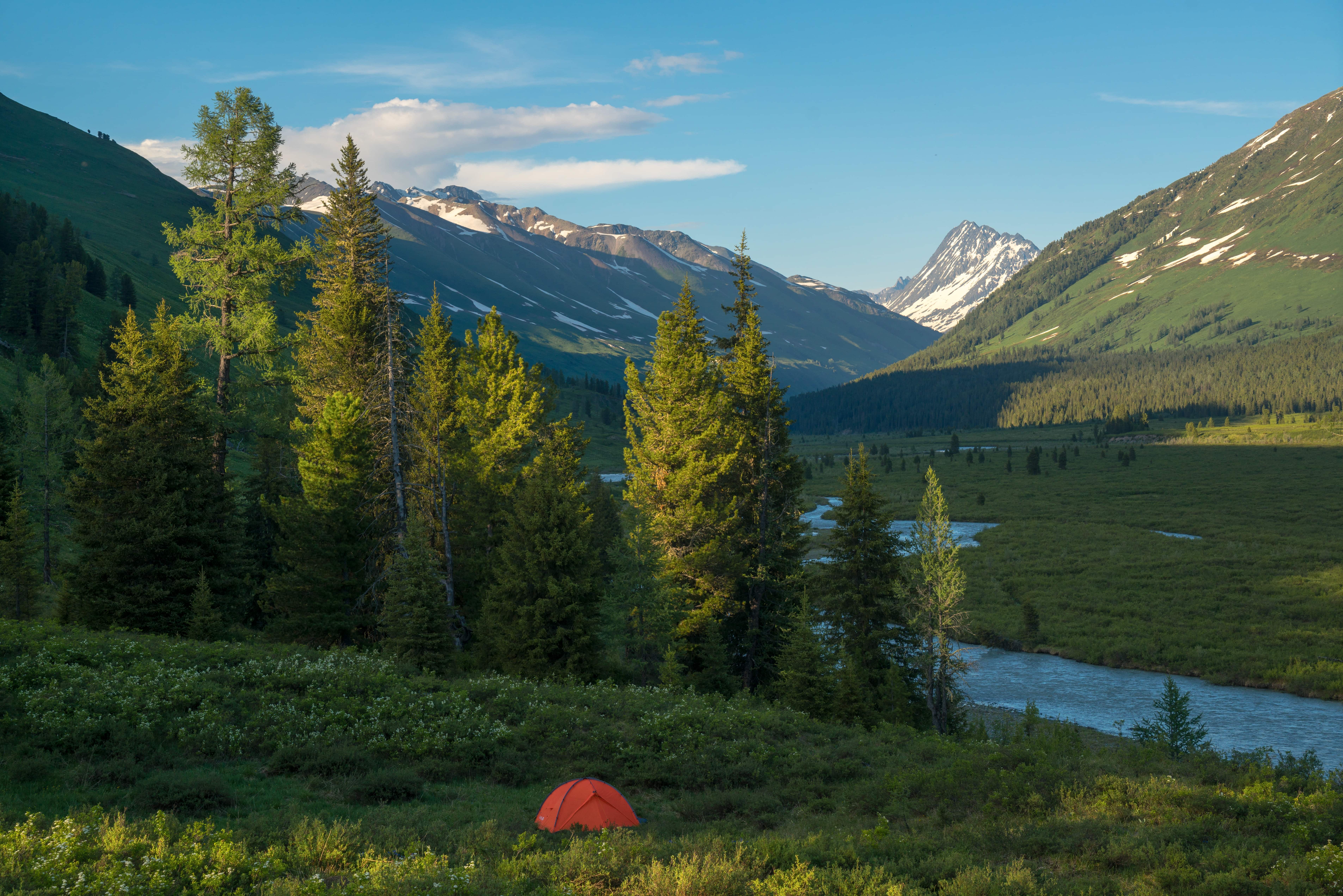 Sunlit valley with evergreen trees, a red tent, a blue lake, and snow-capped mountains.