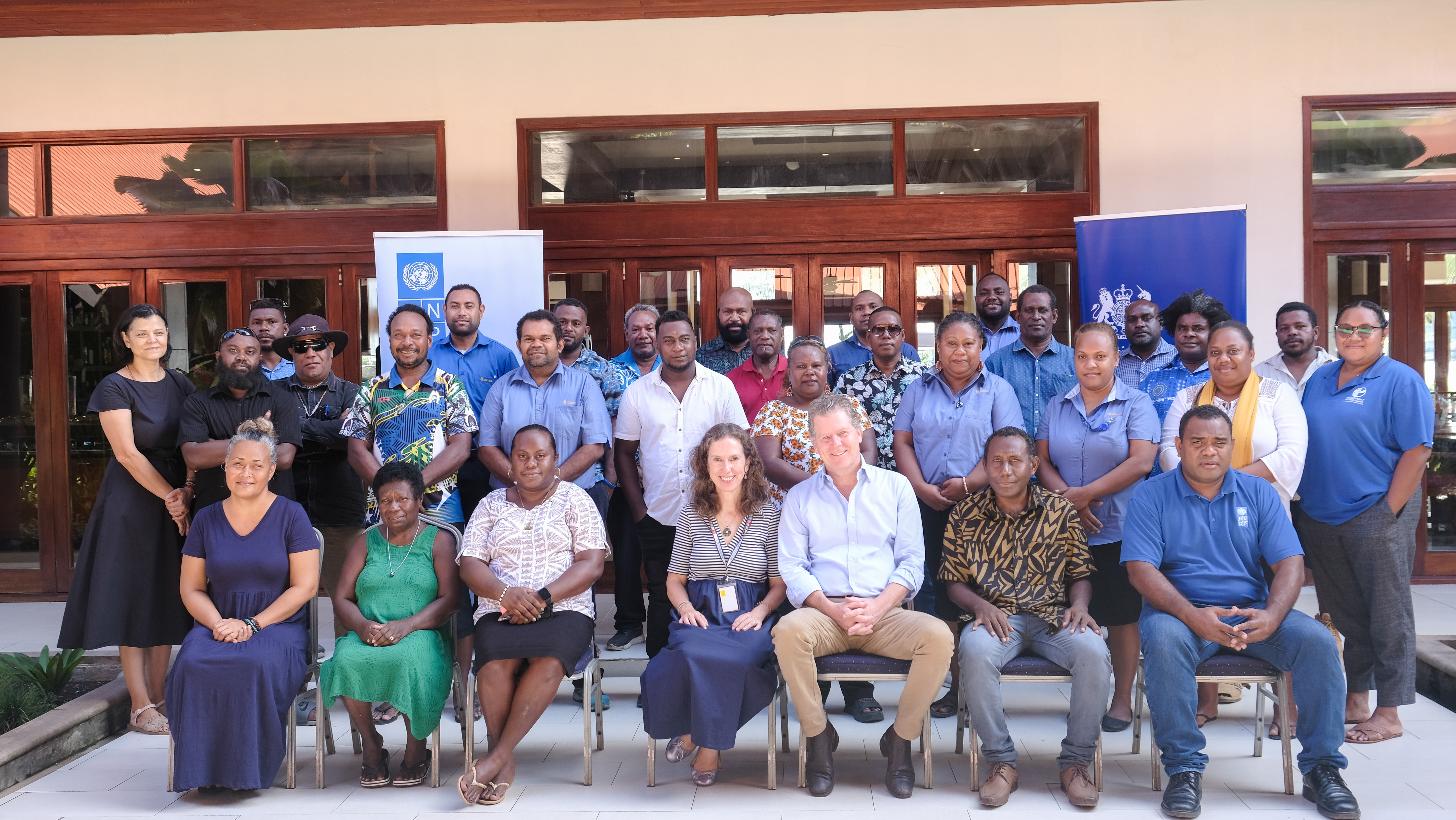 Group photo of diverse adults, standing behind a row of seated people outside a building with blue banners.