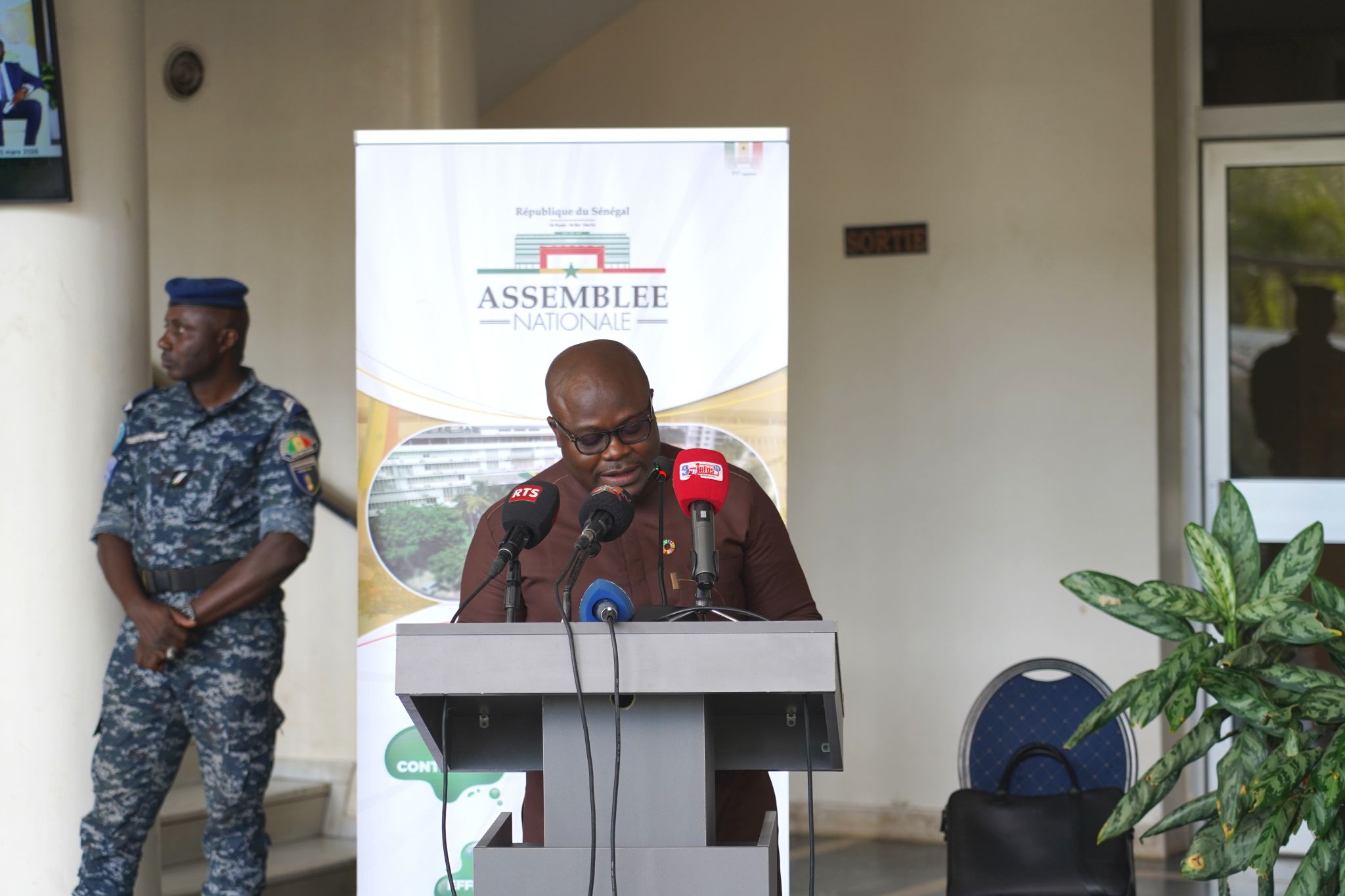 Man at podium speaking, security guard in camouflage to the left, banner and doorway behind.
