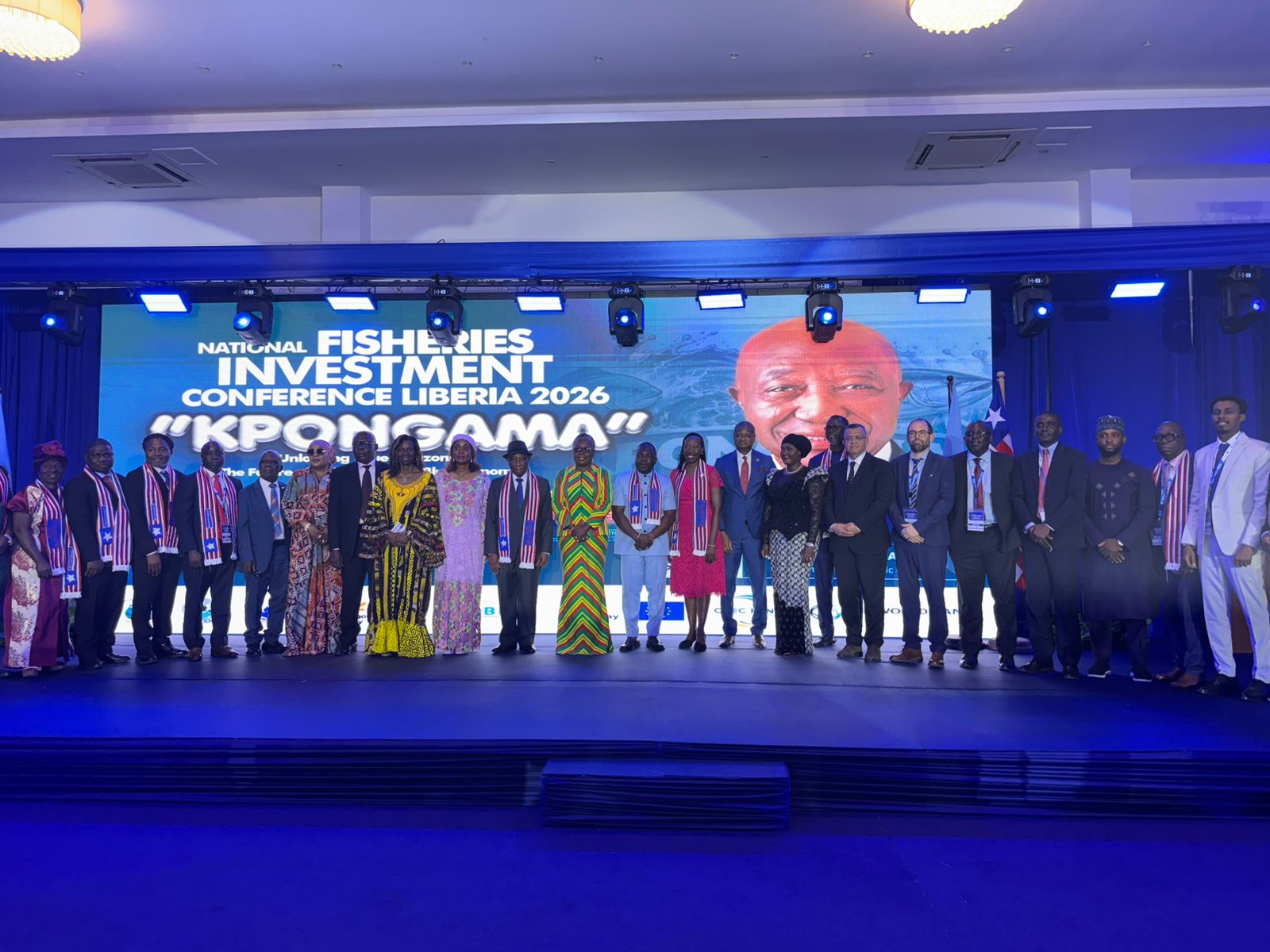 Group of people on a stage posing for a photo at a conference, blue lighting, banner in background.