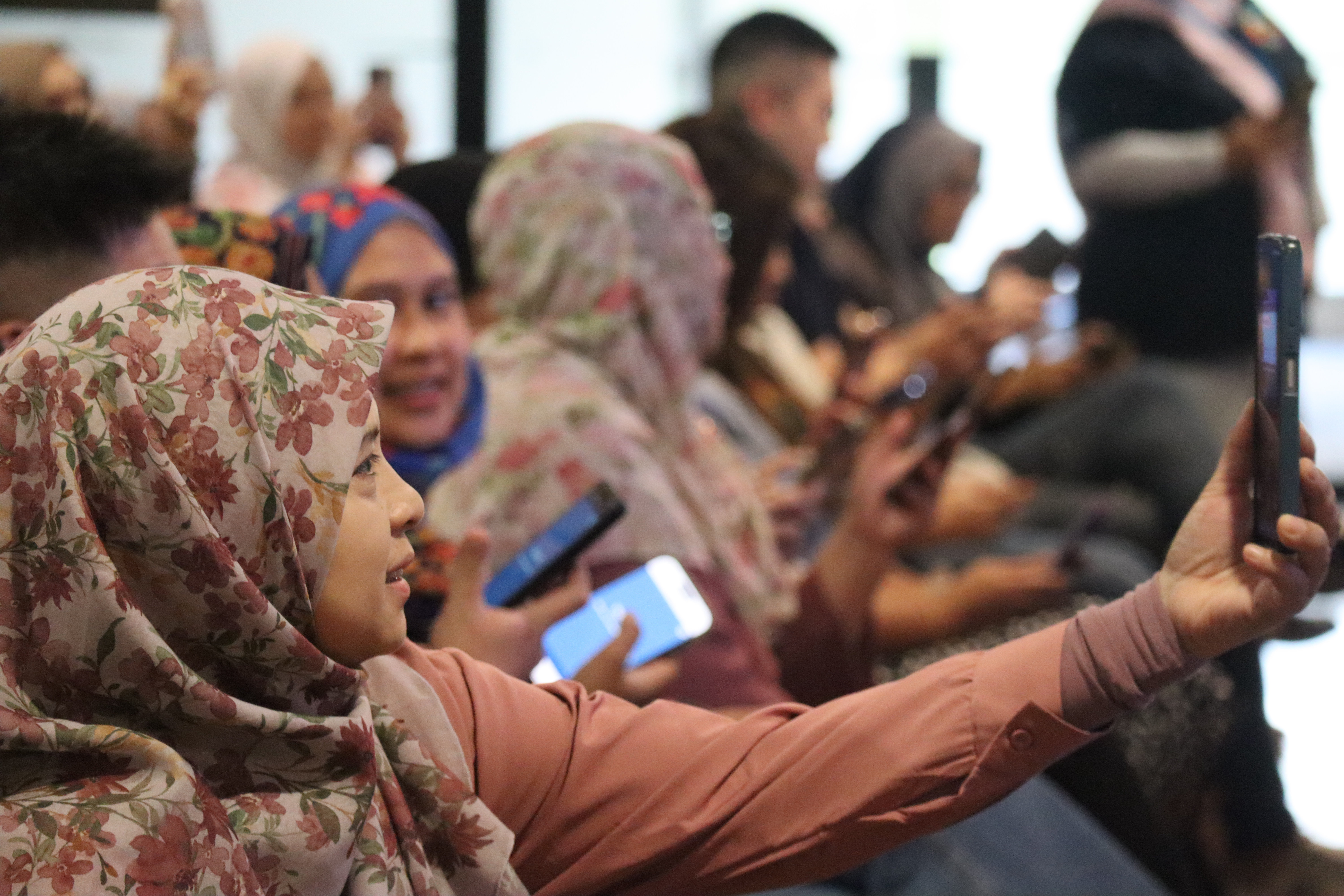 Crowded room of people wearing hijabs seated in a workshop, some using smartphones.