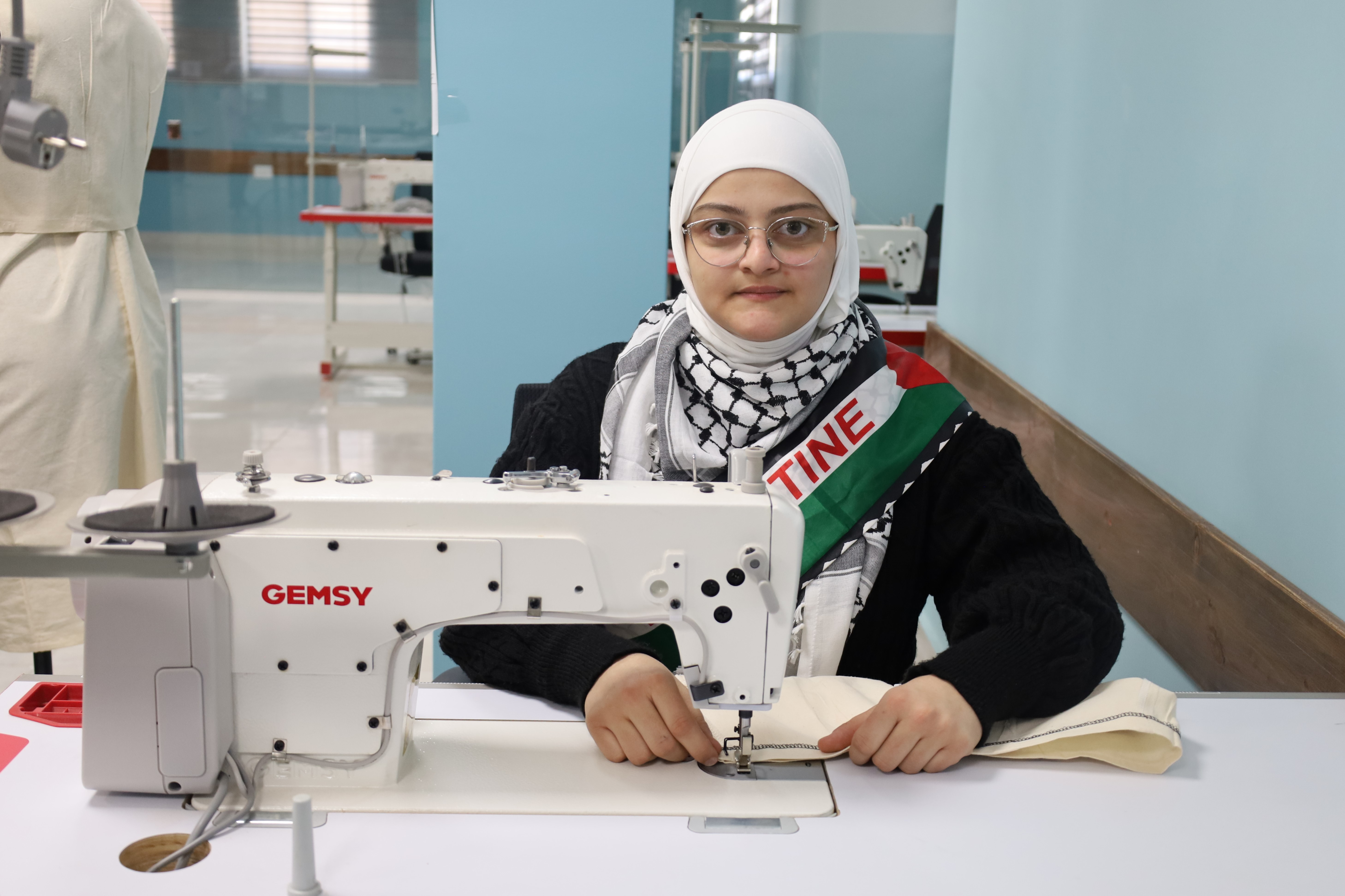 Person sewing at a workstation with a white sewing machine, wearing a checkered scarf.