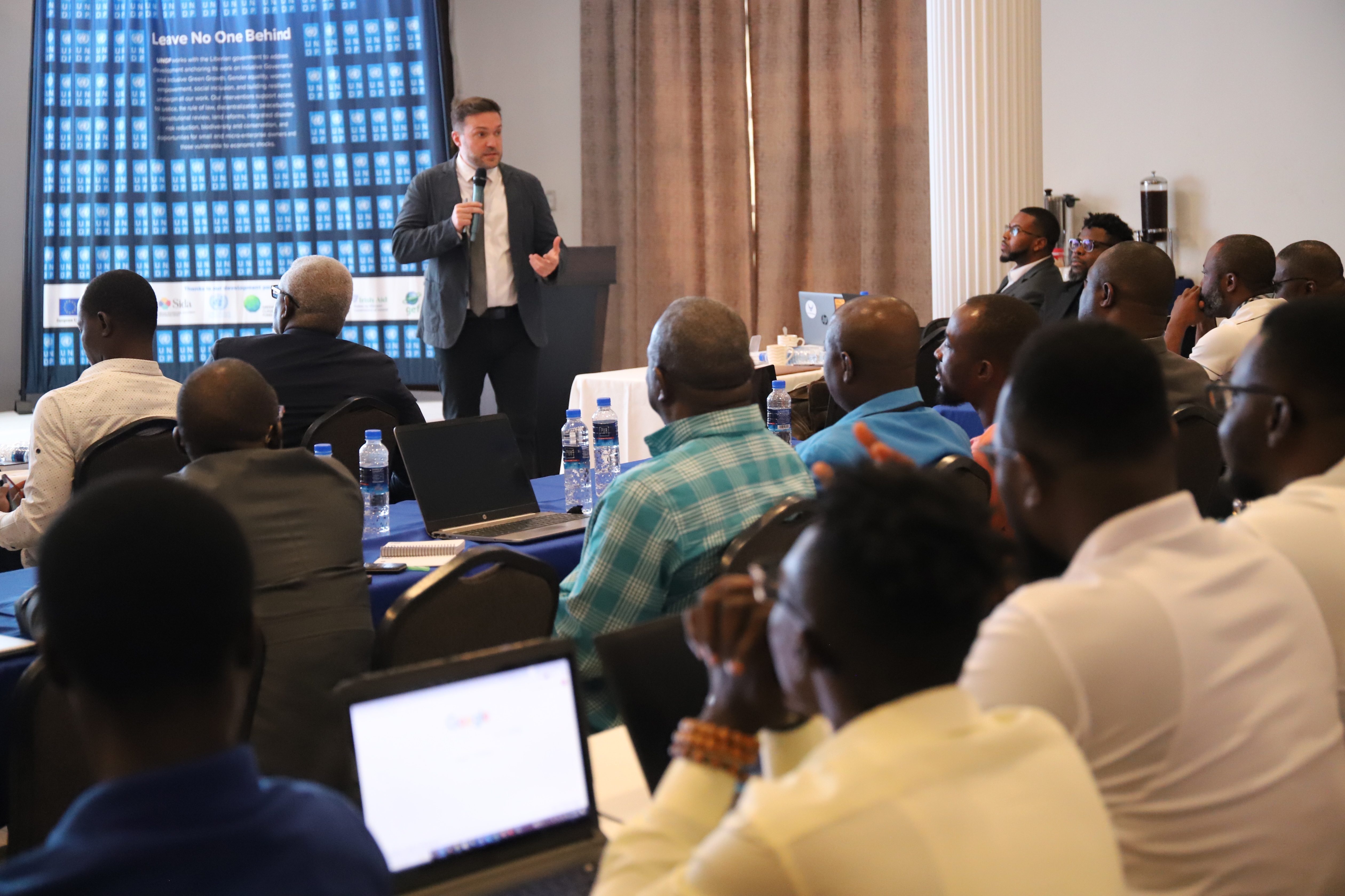 Presenter in a suit at the front of a classroom; audience sits, some with laptops, slide projected.