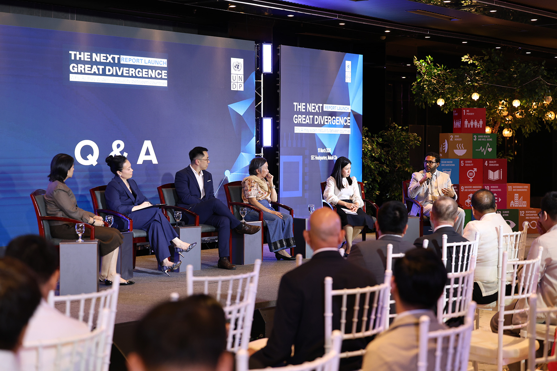 Panel discussion on stage with several speakers at a conference, blue lighting, audience seated.