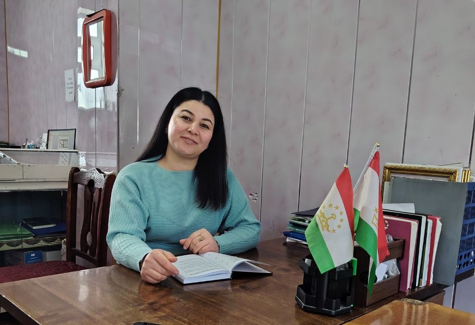 Woman sits at a wooden desk indoors, writing in a notebook with flags on the desk.