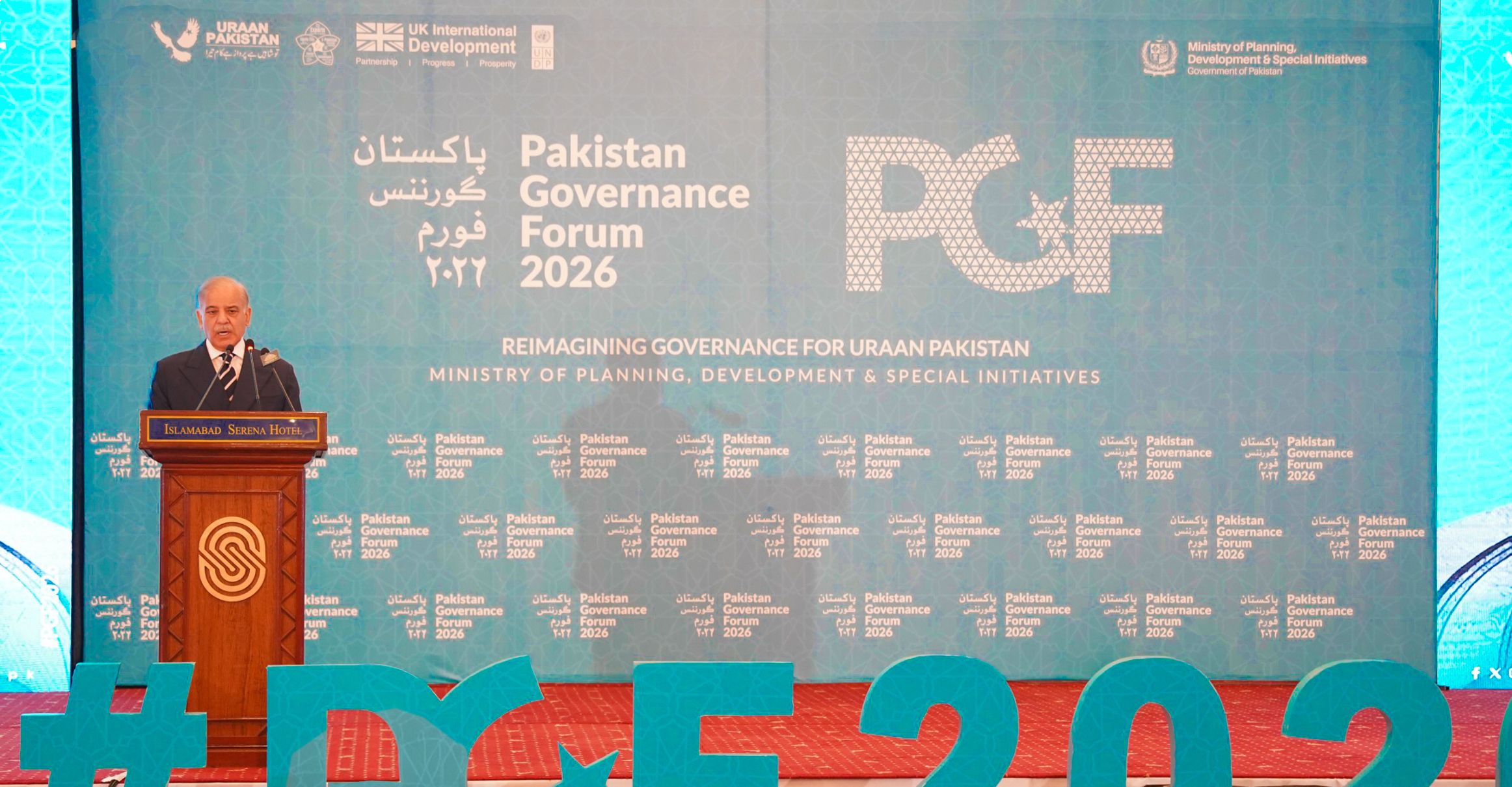 Photograph: man at podium delivering a speech on a conference stage with a blue backdrop and PGF logo.