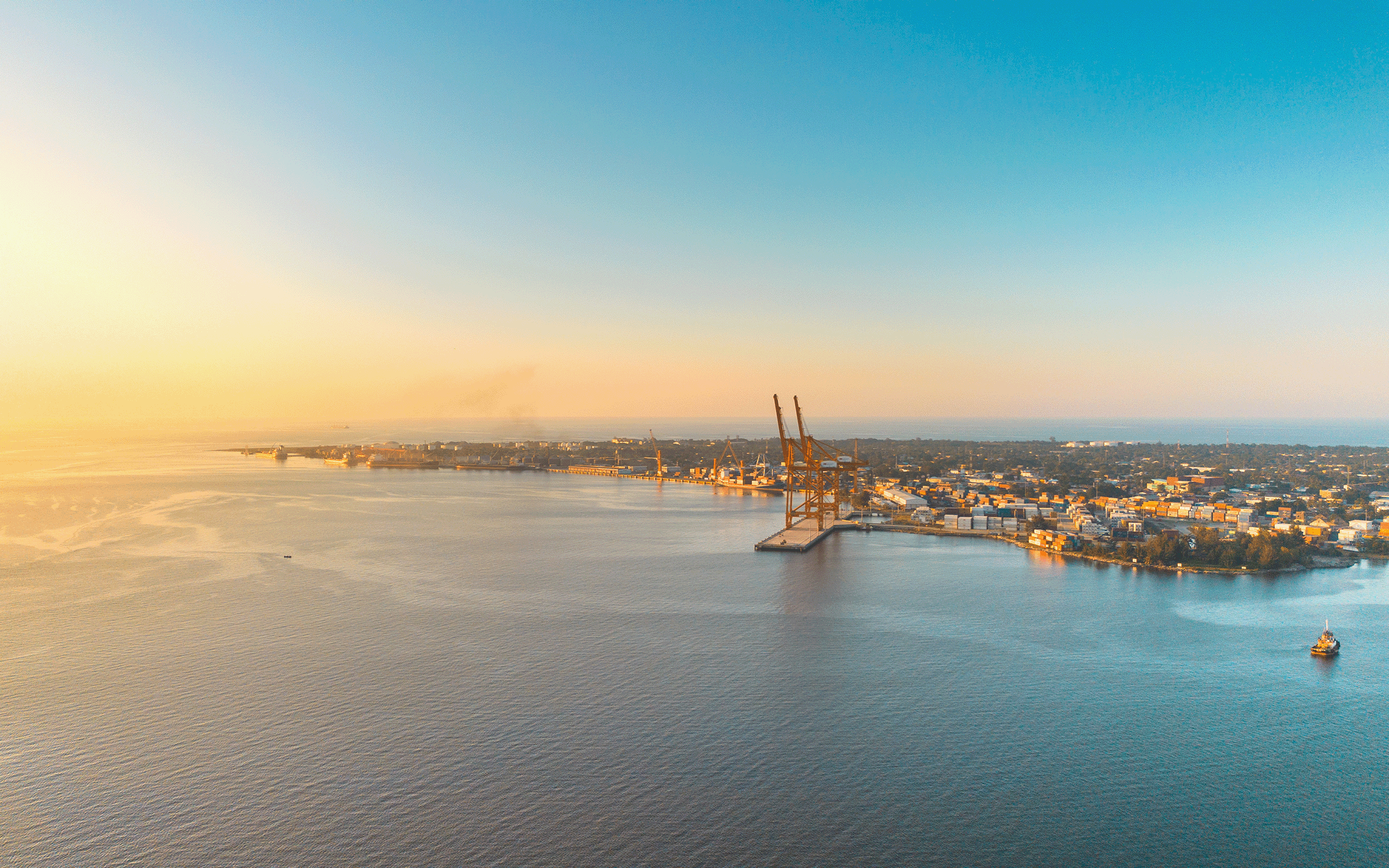 A photograph of a harbor at sunset with cranes and a distant city skyline across calm water.