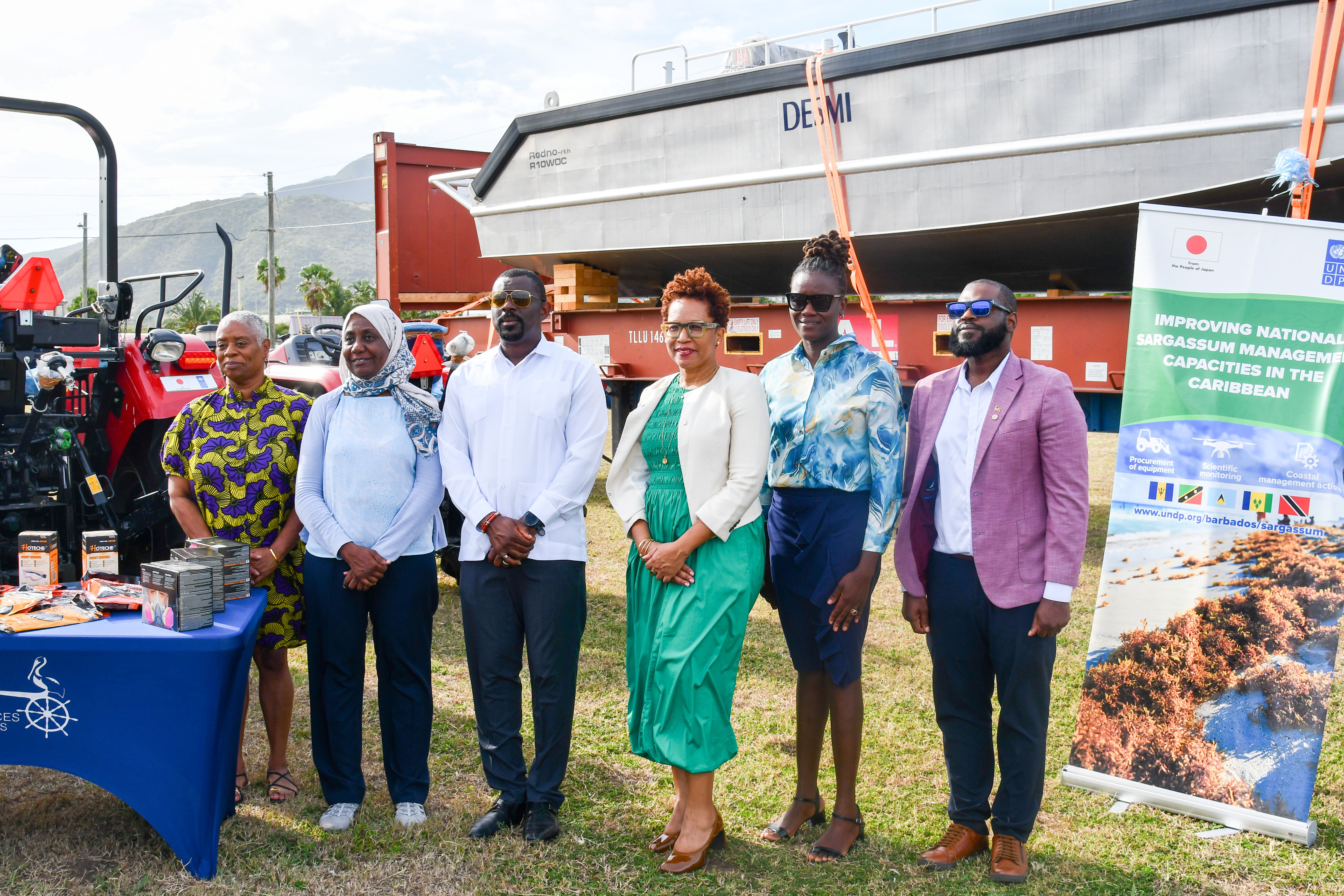 Photo: Group of people standing in a row at an outdoor event, with a blue table and banners.