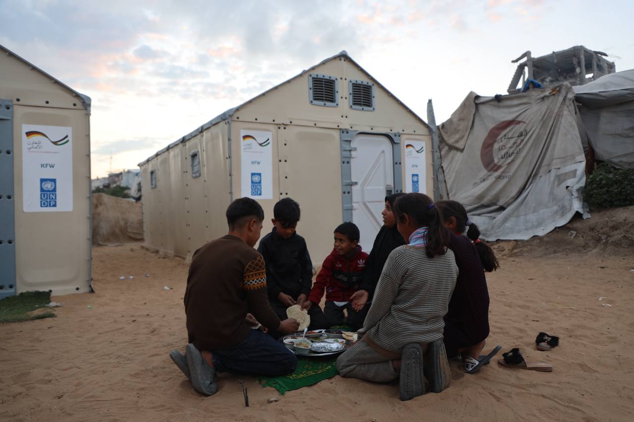 Group of people sits in a circle on sandy ground at a camp, with portable toilets in background.