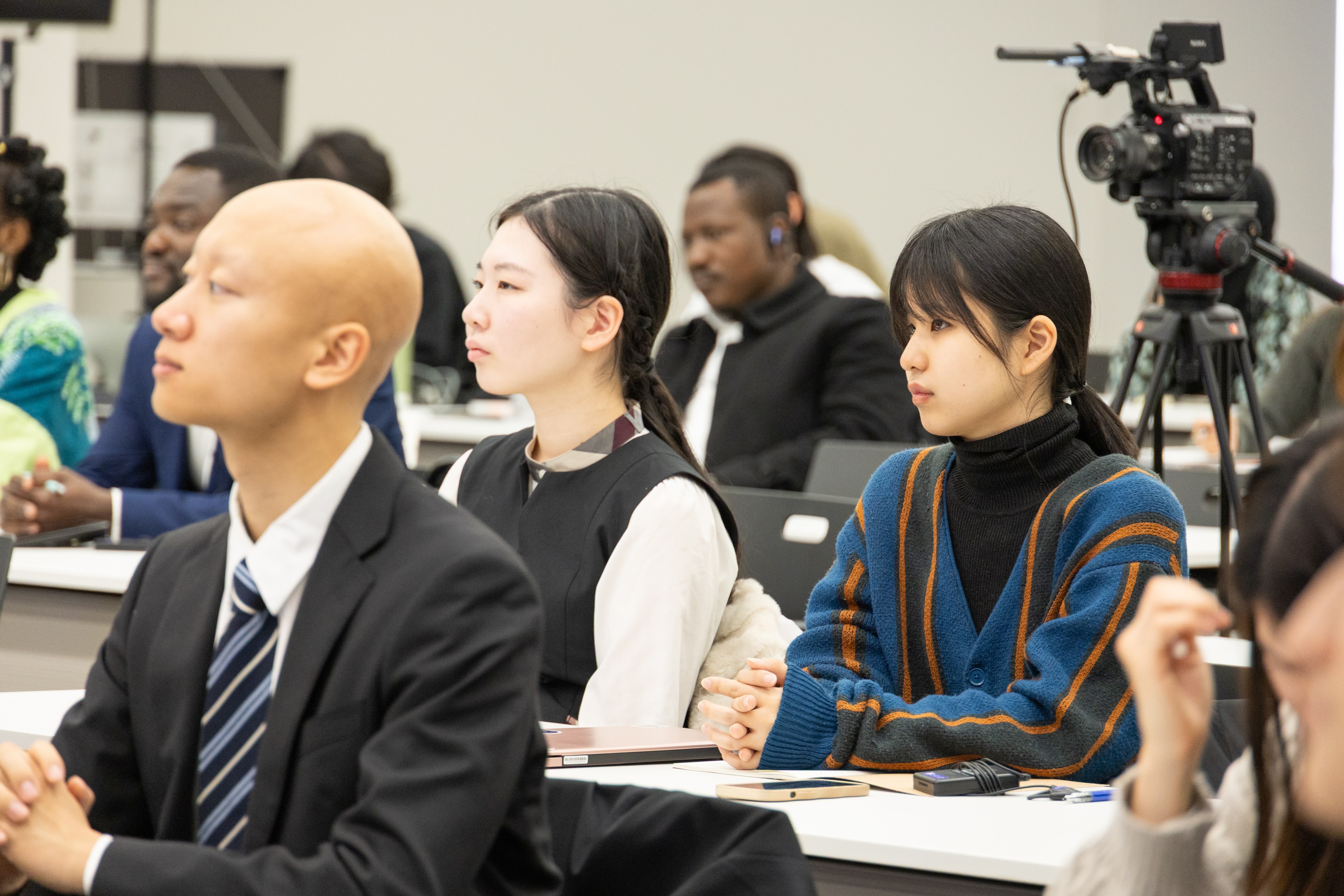 Participants in a conference room sit at desks as a camera crew records.