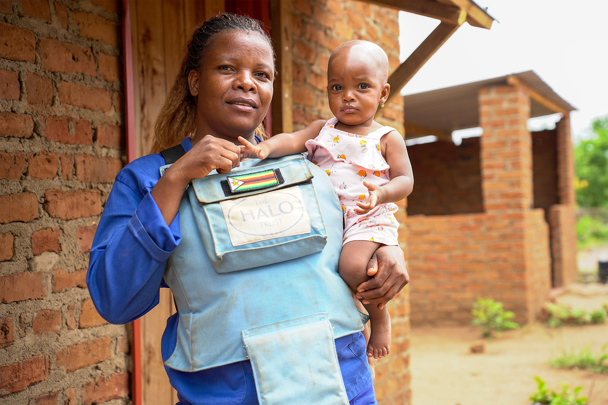 A deminer woman carries a baby in a front carrier outdoors beside a brick building.