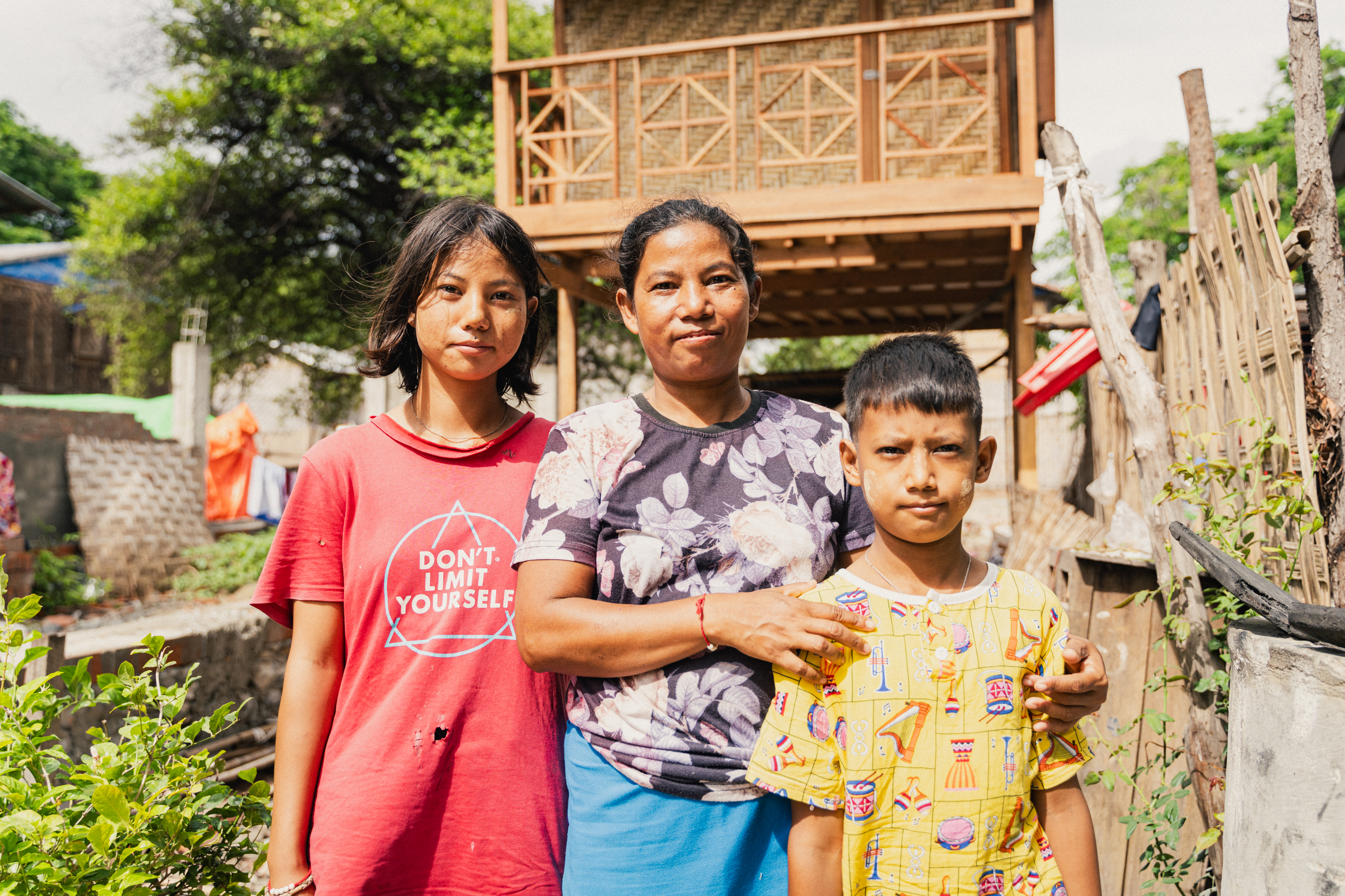 Two children and their mother in colorful shirts stand together in front of a wooden house in a village.