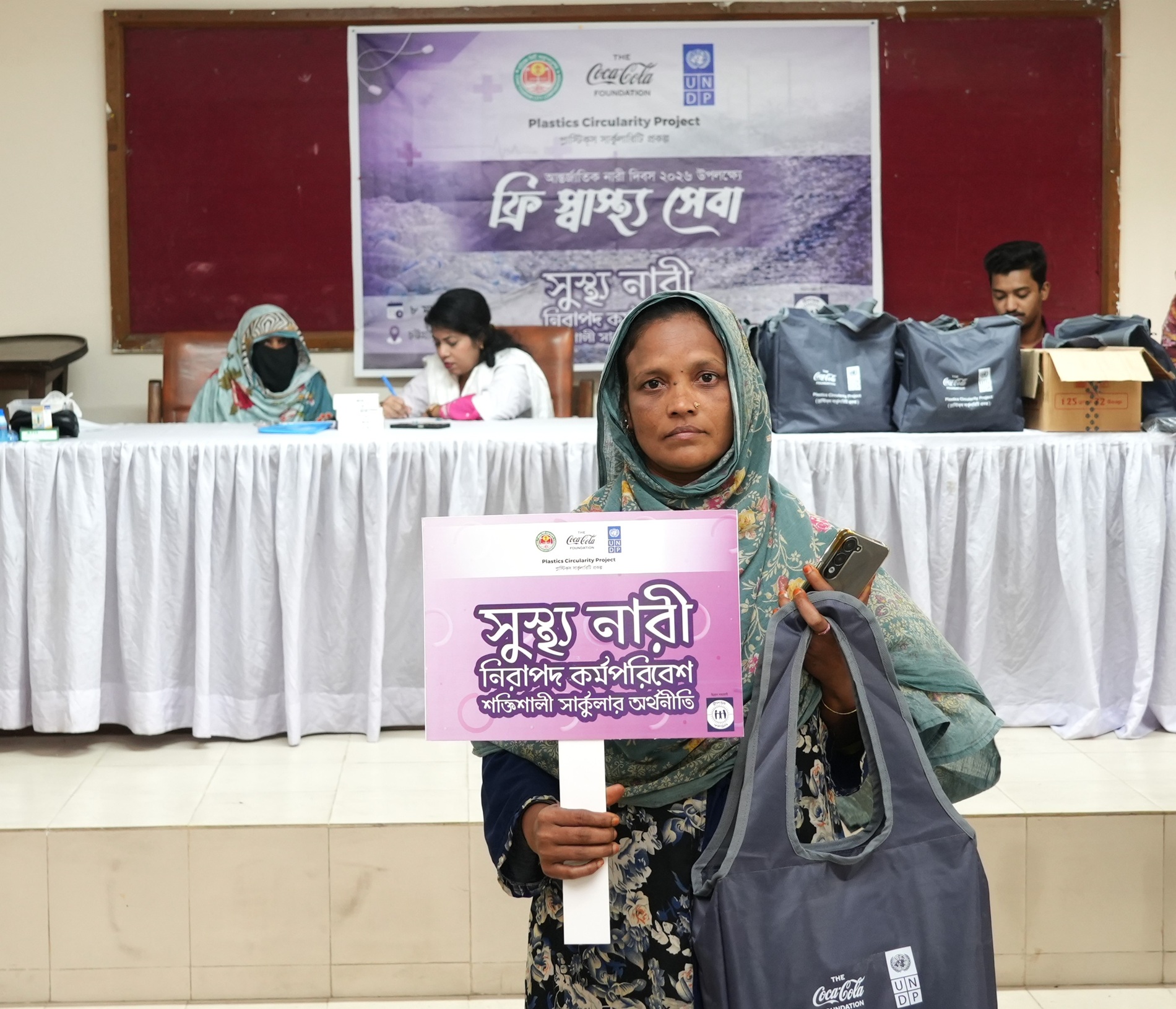 Person wearing a green hijab holds a sign on a stage in front of a panel at a conference.