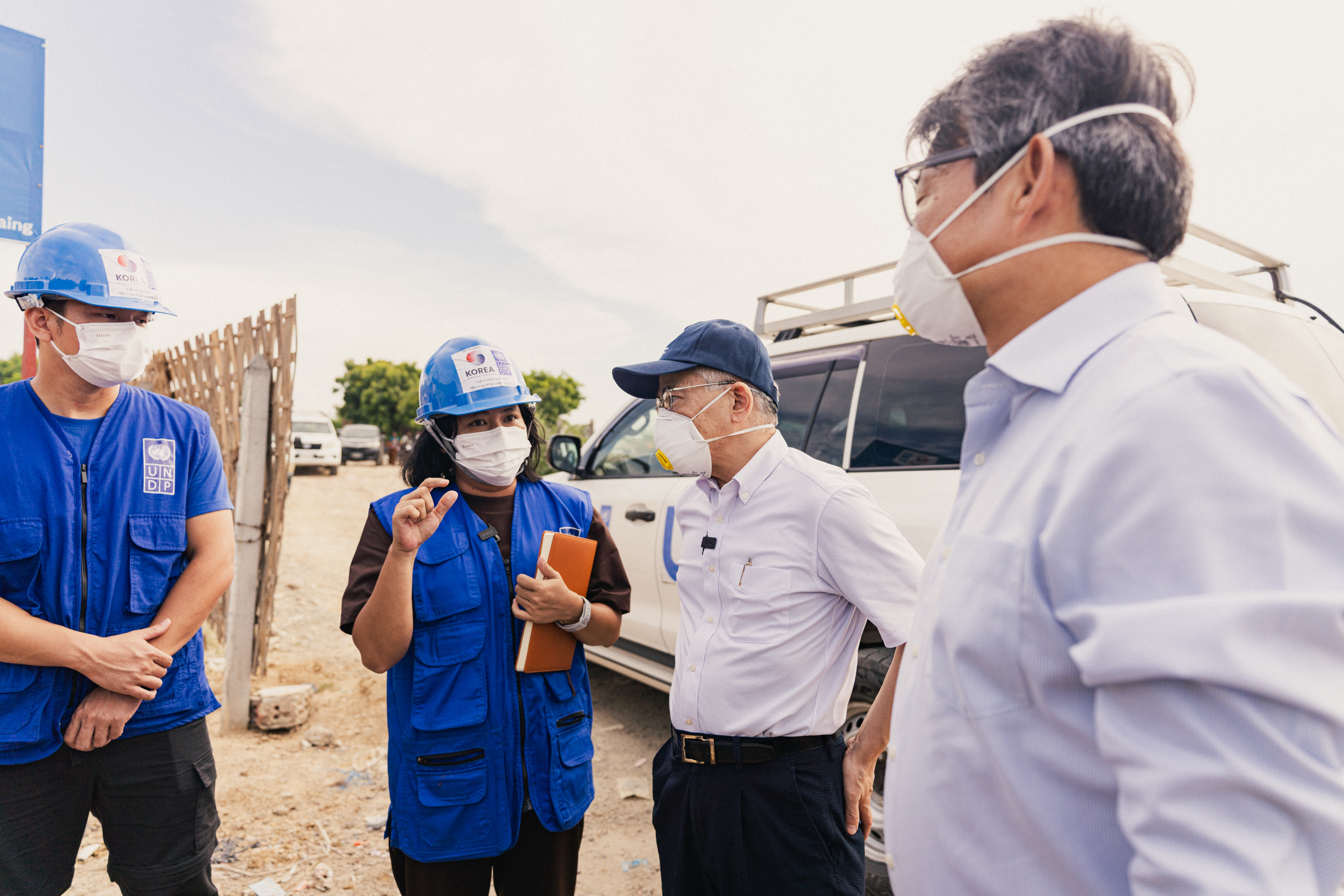 Supervisor wearing a mask talks to workers in blue uniforms and hard hats at a construction site.