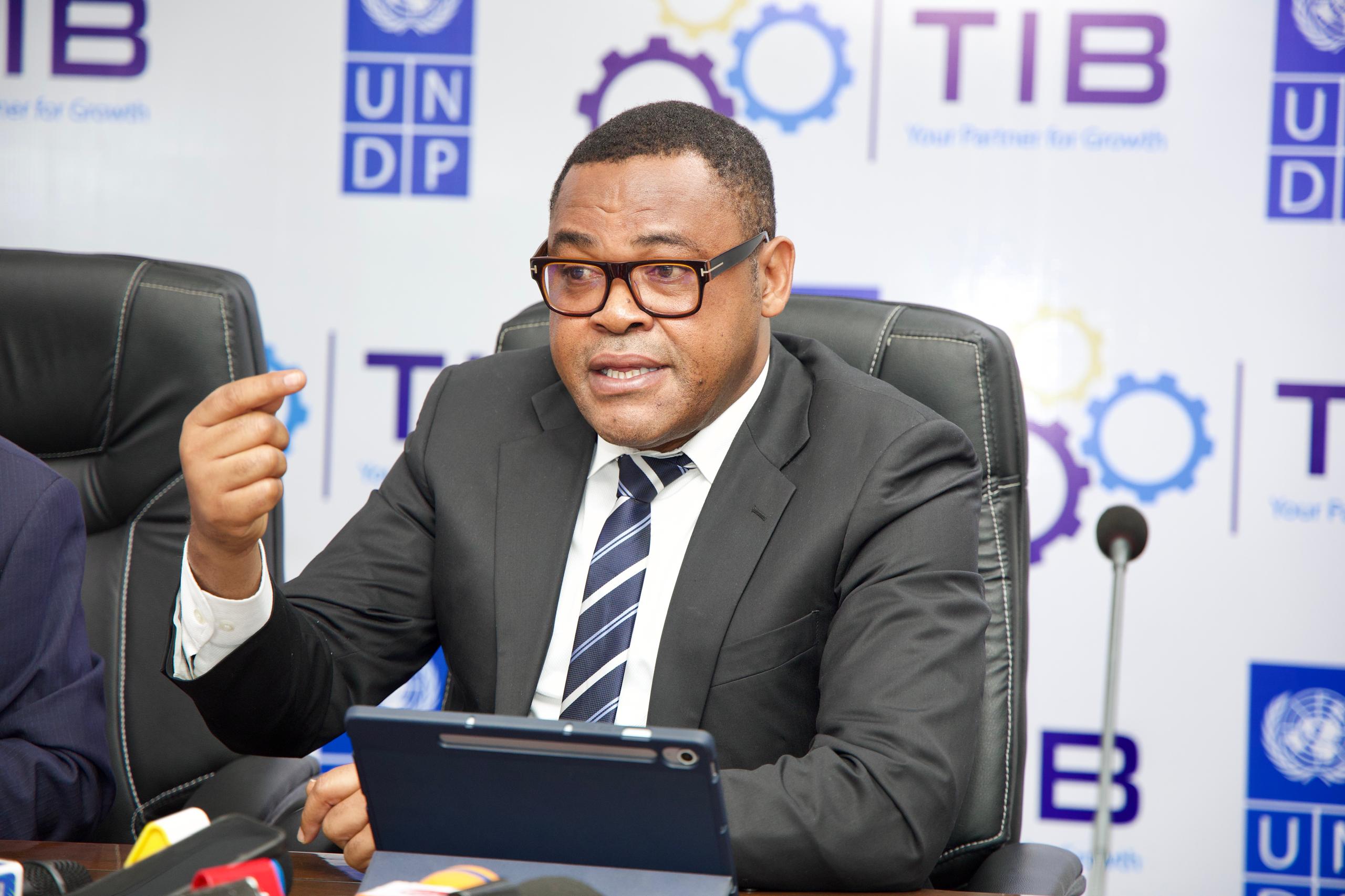 Man in suit with blurred face gestures at a tablet during a meeting; banners behind.