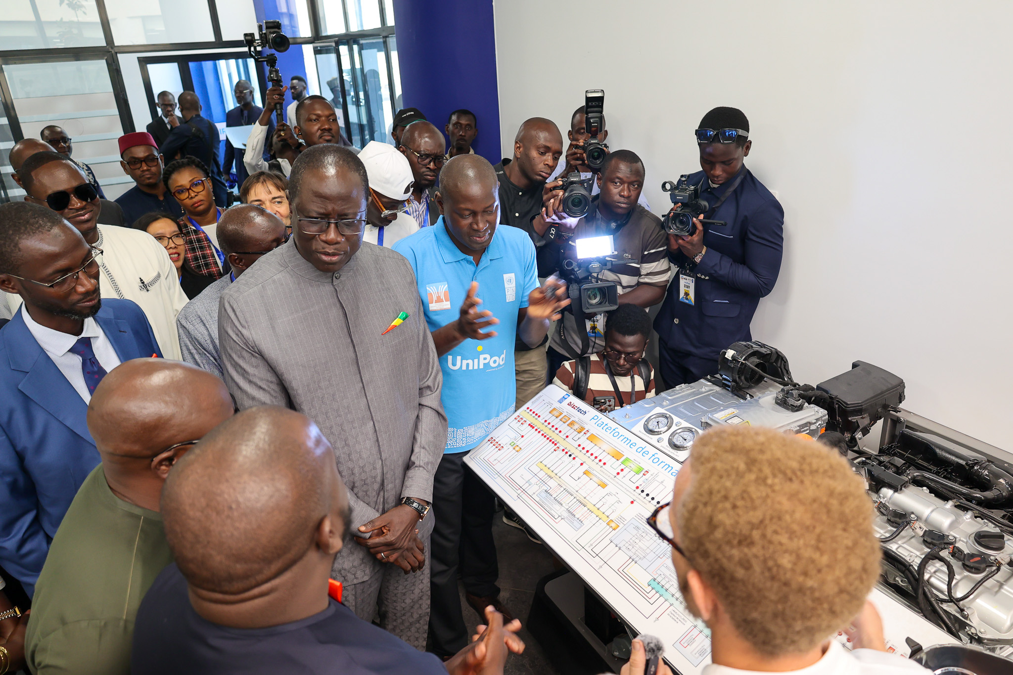 Photo d'une foule rassemblée autour d'une table sur laquelle sont disposés des appareils électroniques, tandis qu'un homme en costume gris prend la parole.