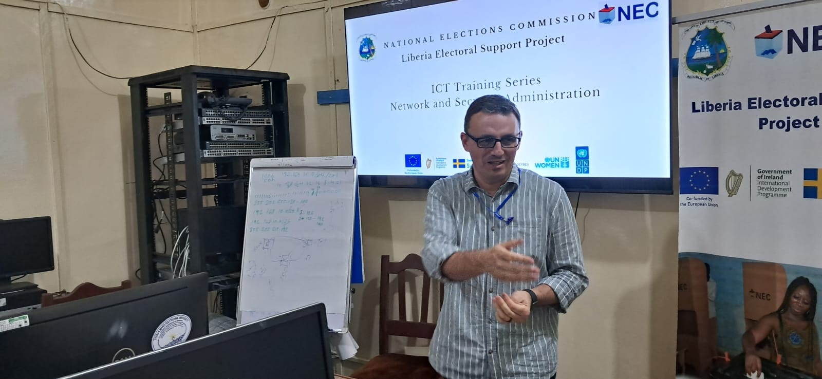 Man in blue shirt presenting in a conference room with projector screen and banners with logos.