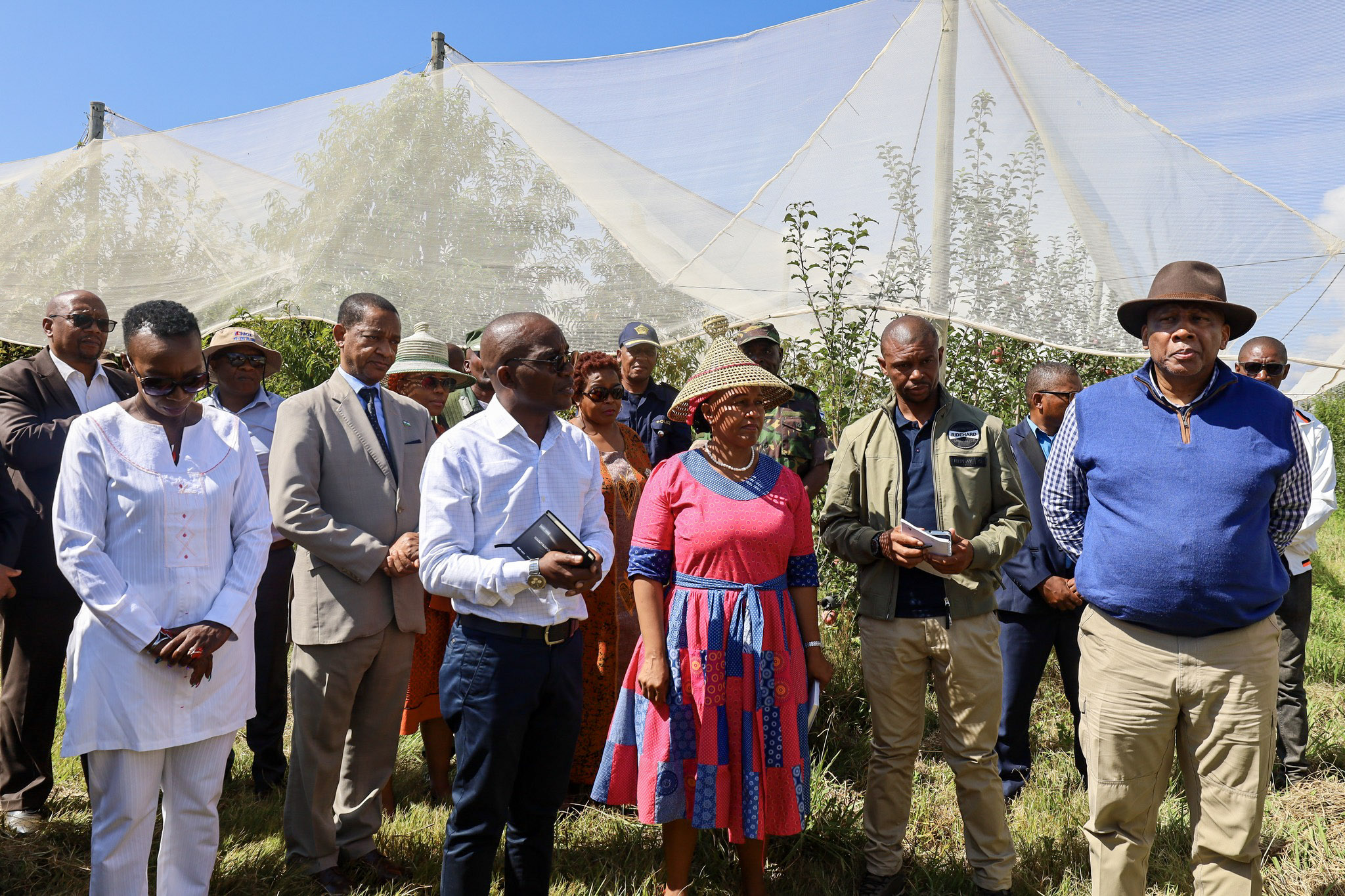 Group photo of people posing outdoors in an orchard with white shade nets protecting apple trees in the background.
