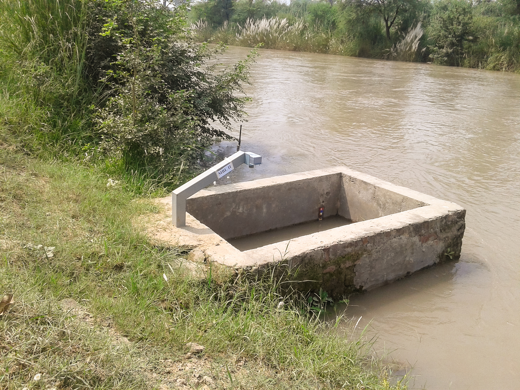 Rectangular concrete trough on a grassy riverbank beside muddy brown water.