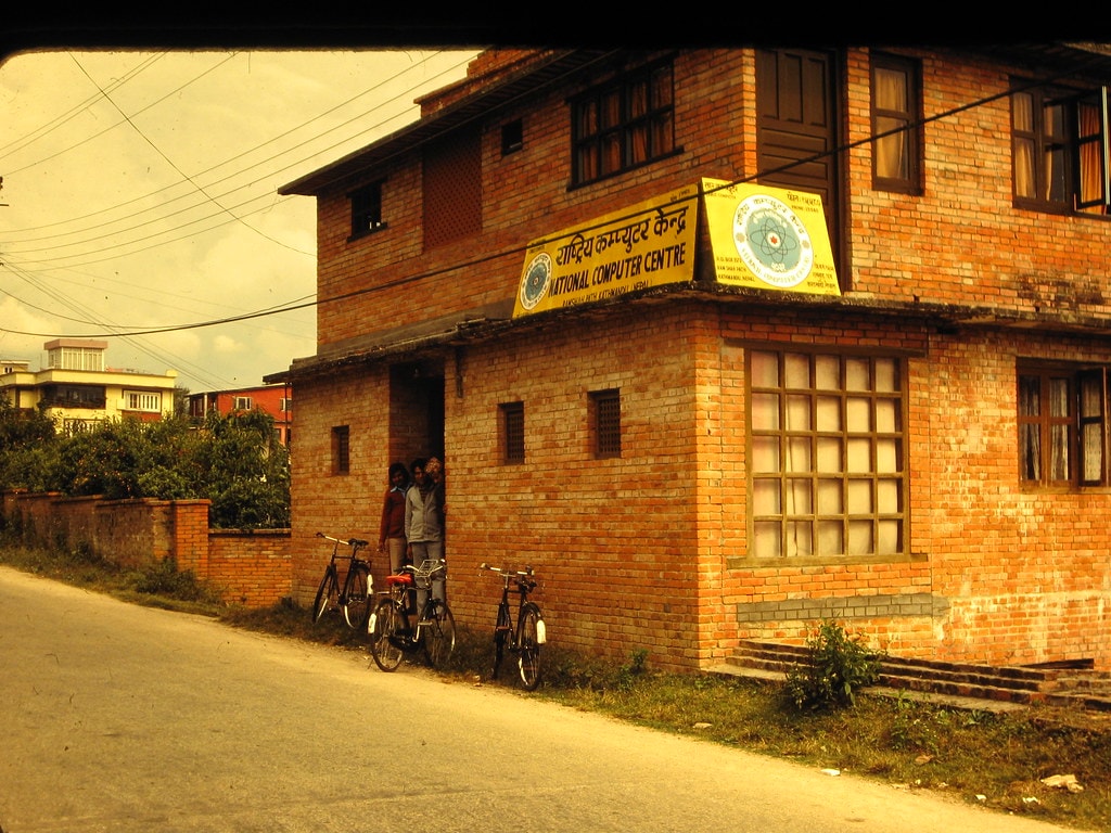 Sunlit two-story brick building with signs; bicycles parked along a dusty street.