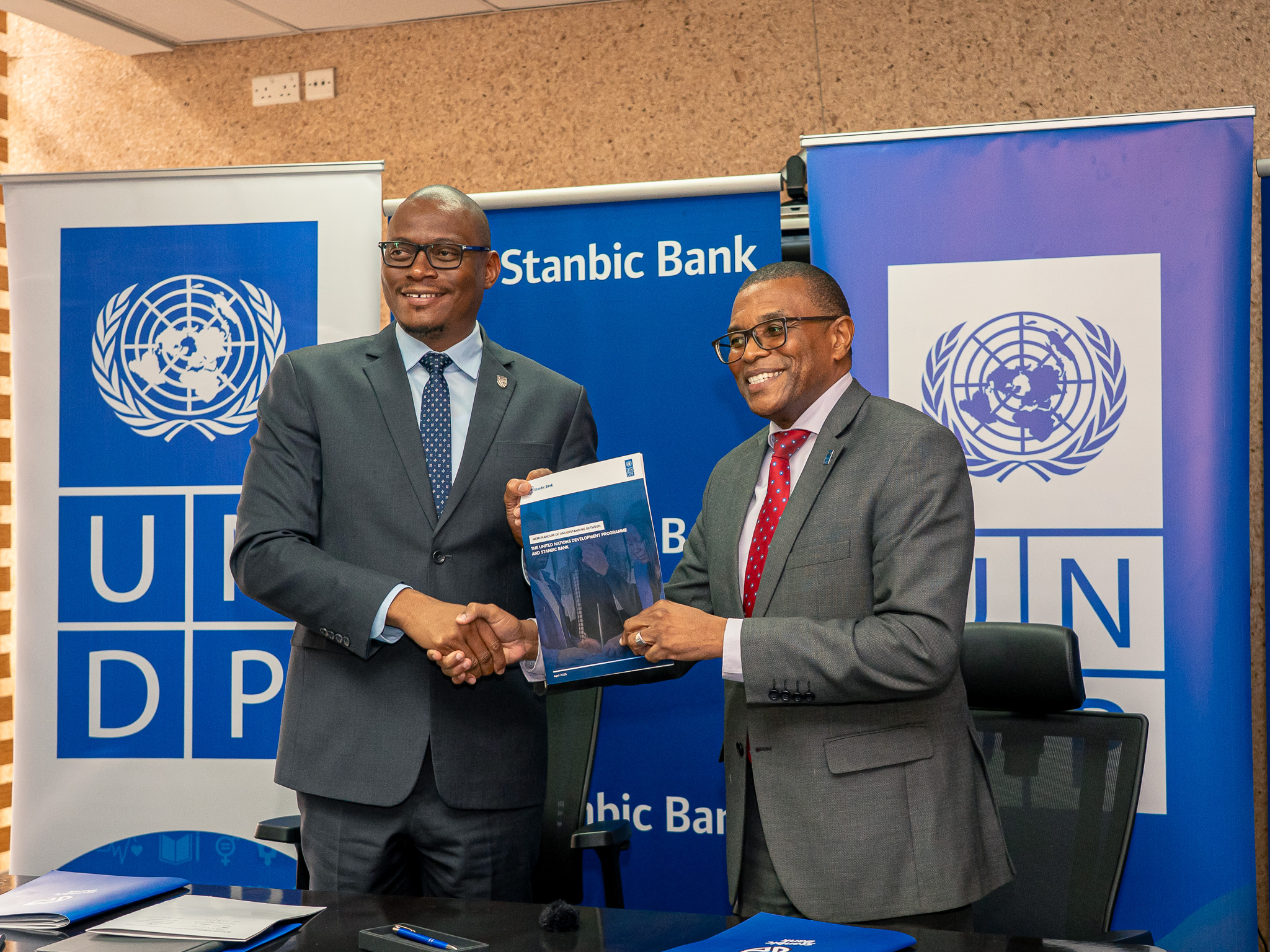 Two men in suits shake hands while exchanging a folder in front of UN banners.