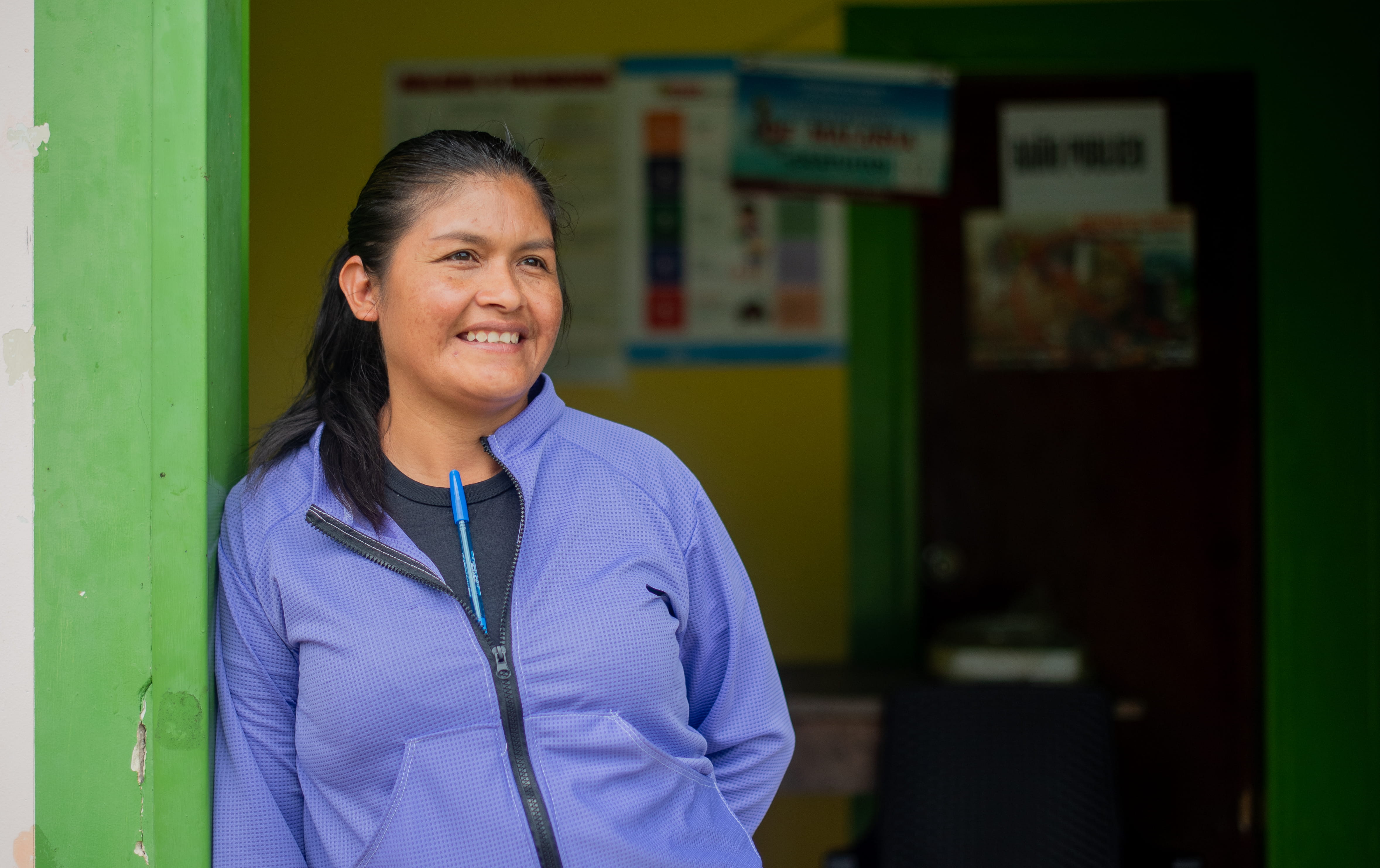 Photograph of a person in a blue jacket standing in a doorway; green wall and posters in the background, face blurred.