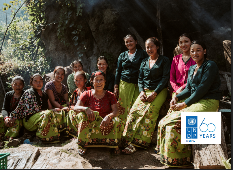 Group of smiling people in colorful traditional outfits sitting outdoors under a tree.