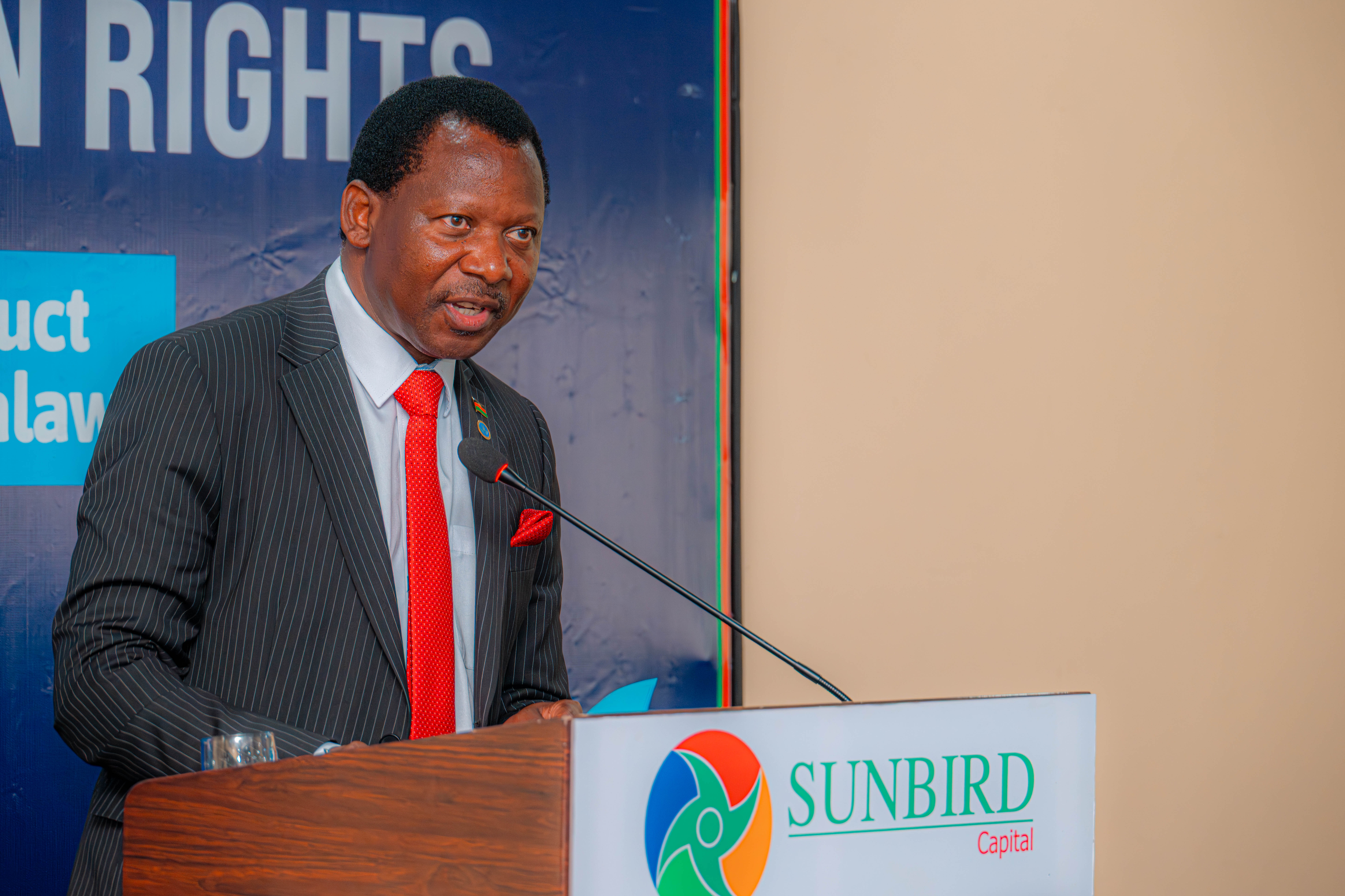 Man in suit with red tie speaks at a lectern bearing a Sunbird logo, with a blue backdrop.