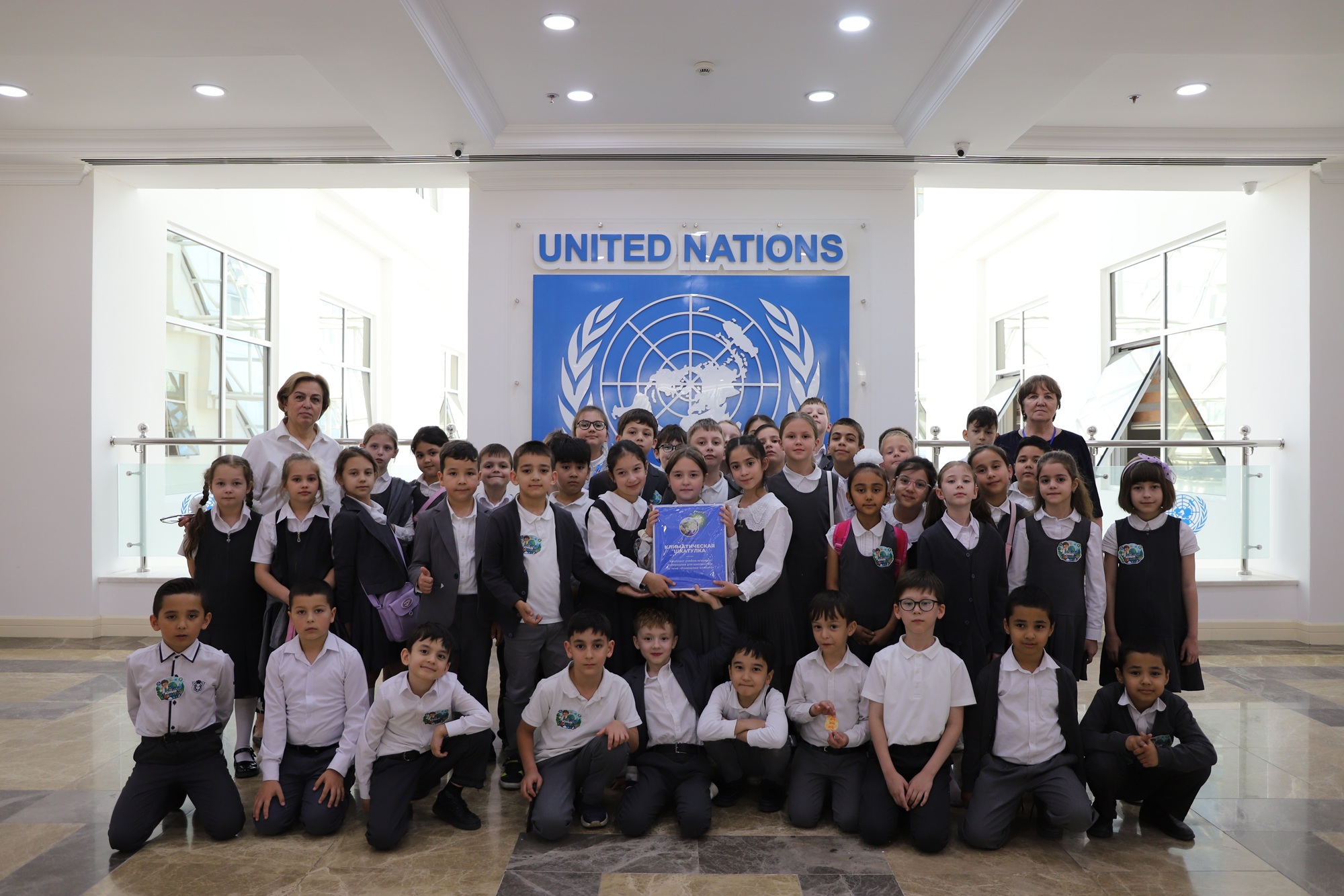 Group photo of students in formal attire posing in front of a United Nations banner in a bright hall.