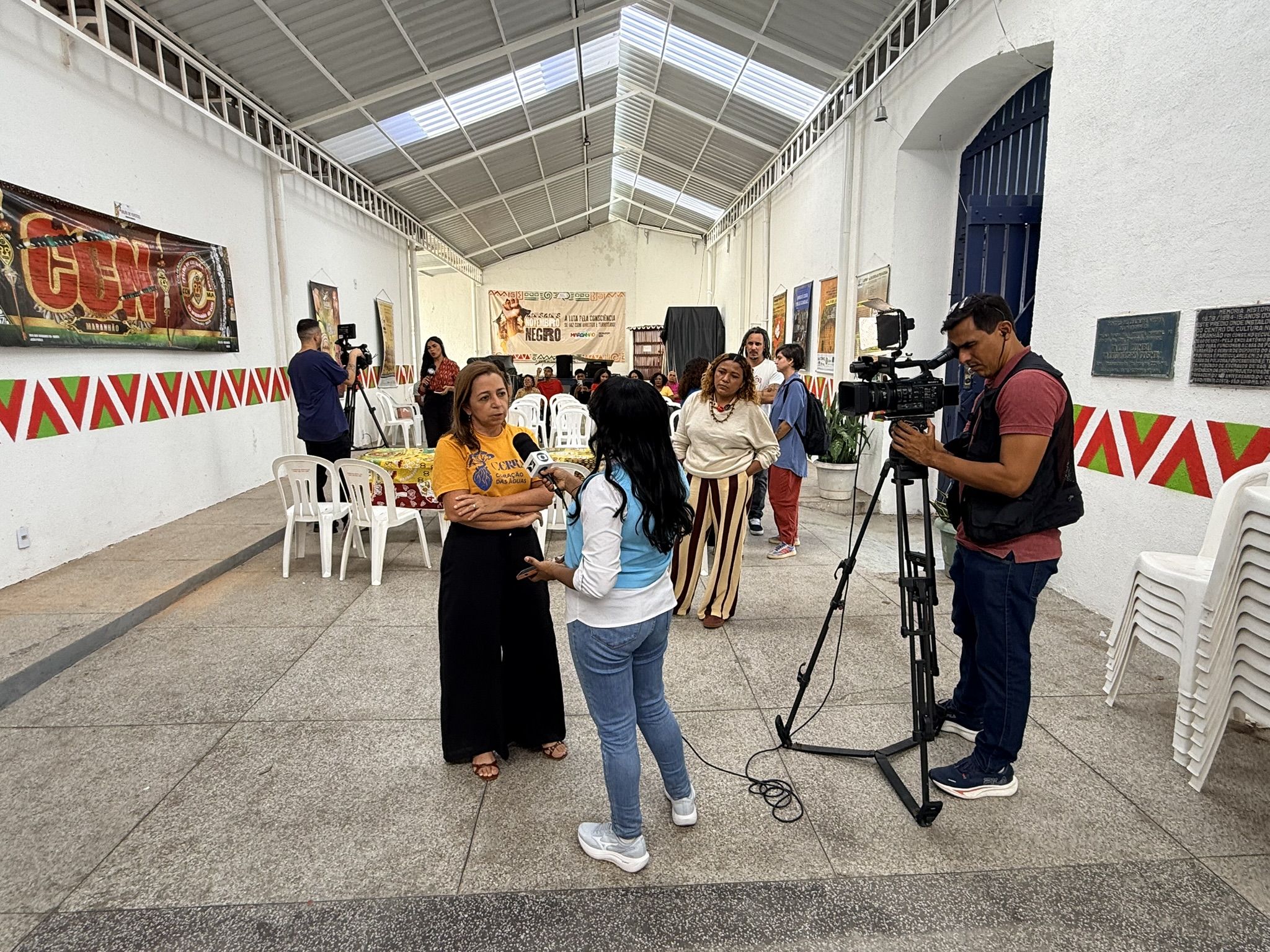 Group of people in a bright hallway with red chevron trim; camera crew films a young woman.