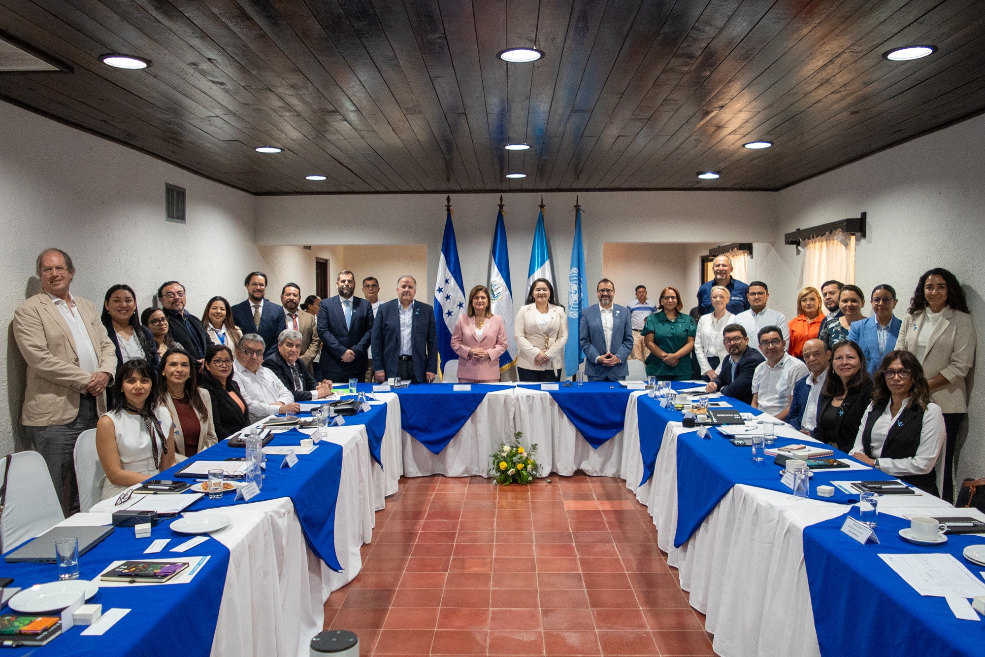 Group of professionals around a blue-and-white U-shaped conference table with flags in the background.