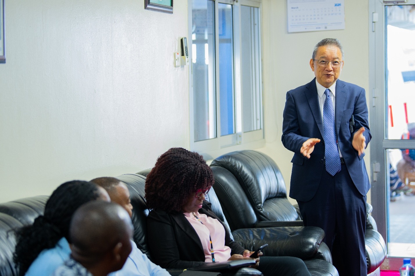 Photograph: man in a suit speaks to a small group seated on a couch in a bright room.