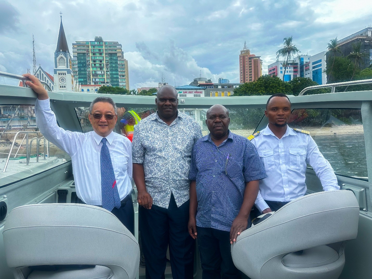Four men pose on a boat deck with water and a city skyline behind them.