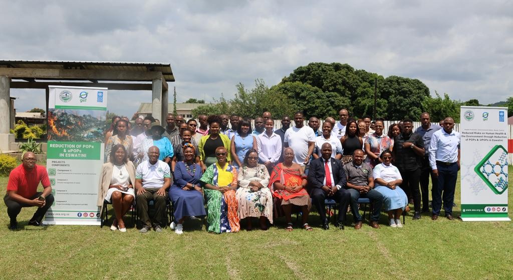 Group photo of diverse people posing outdoors on a grassy field with banners in the background.