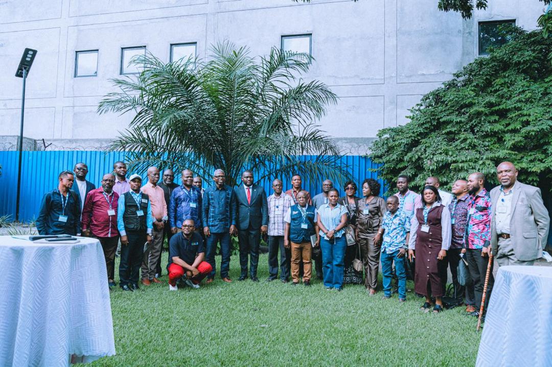 Group of people posing outdoors on a lawn with a palm tree and blue fence.