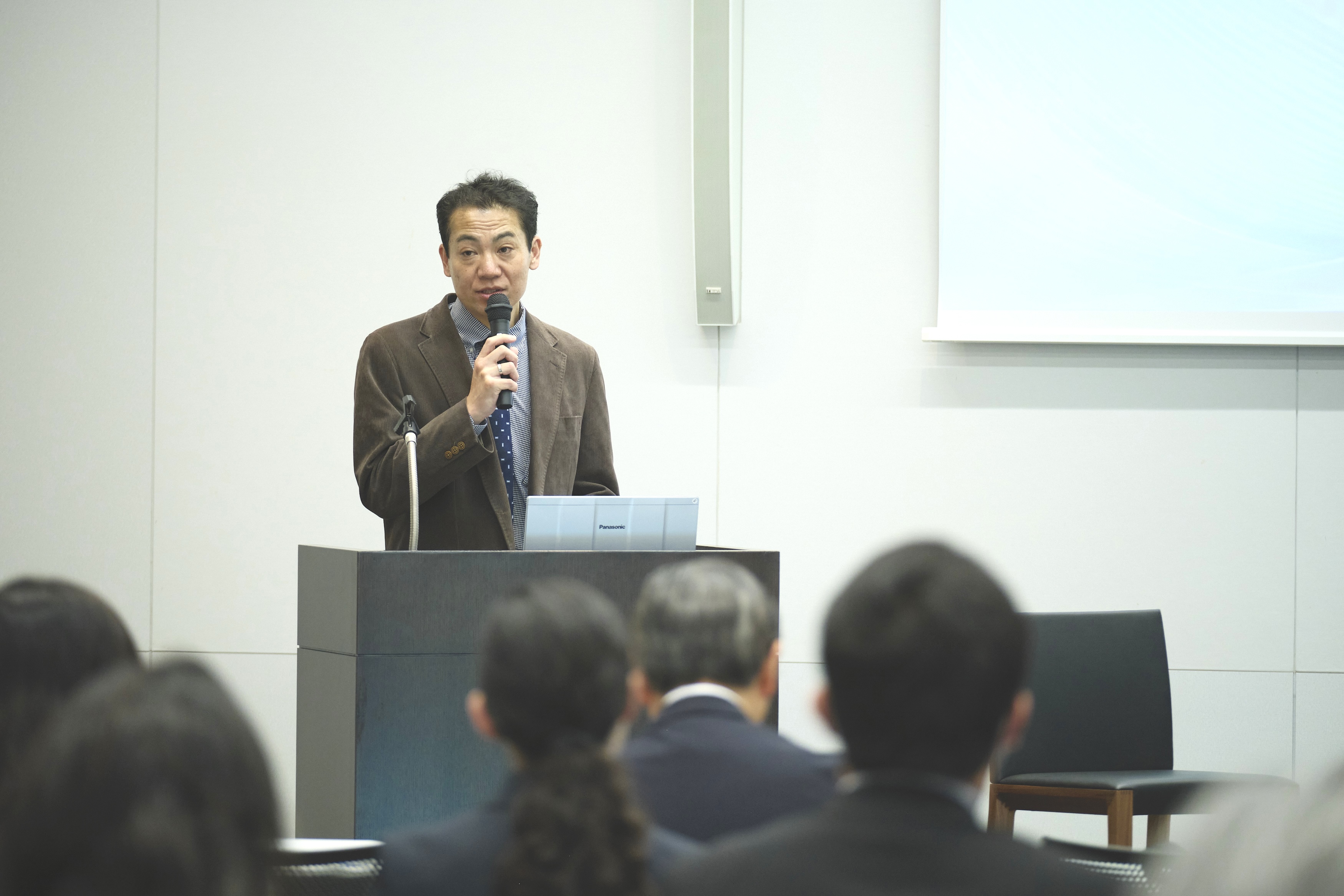 Photograph of a man in a brown blazer speaking at a podium to an audience.