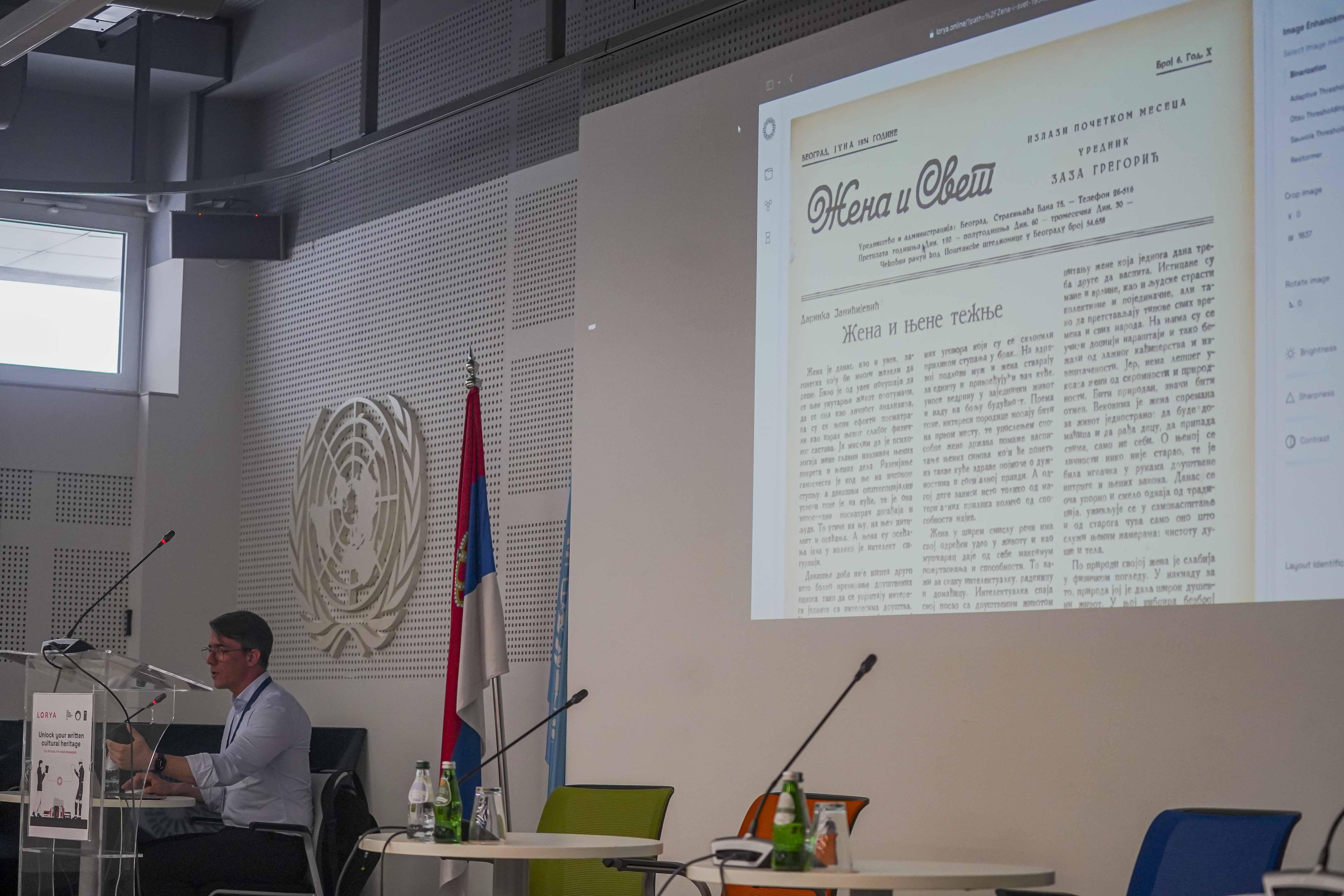 Photograph of a presenter at a conference; projection shows a document, flag and emblem visible.