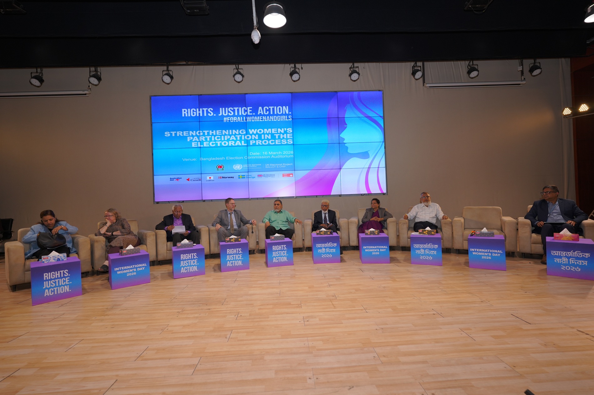 Panel of speakers with name placards on stage at a conference hall, large blue screen behind.
