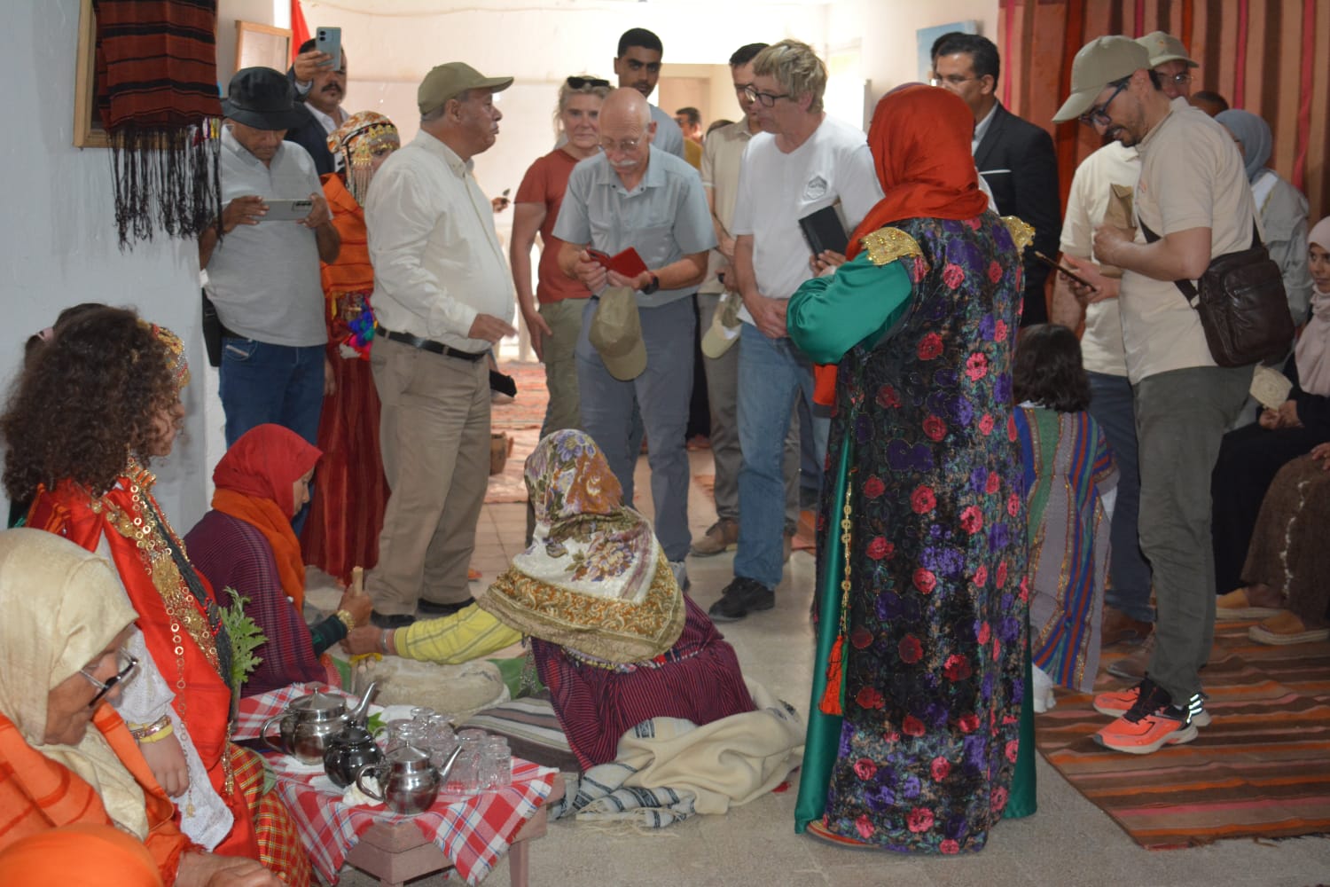 Photo: group in colorful traditional clothes gathers around a person seated on a rug with baskets.