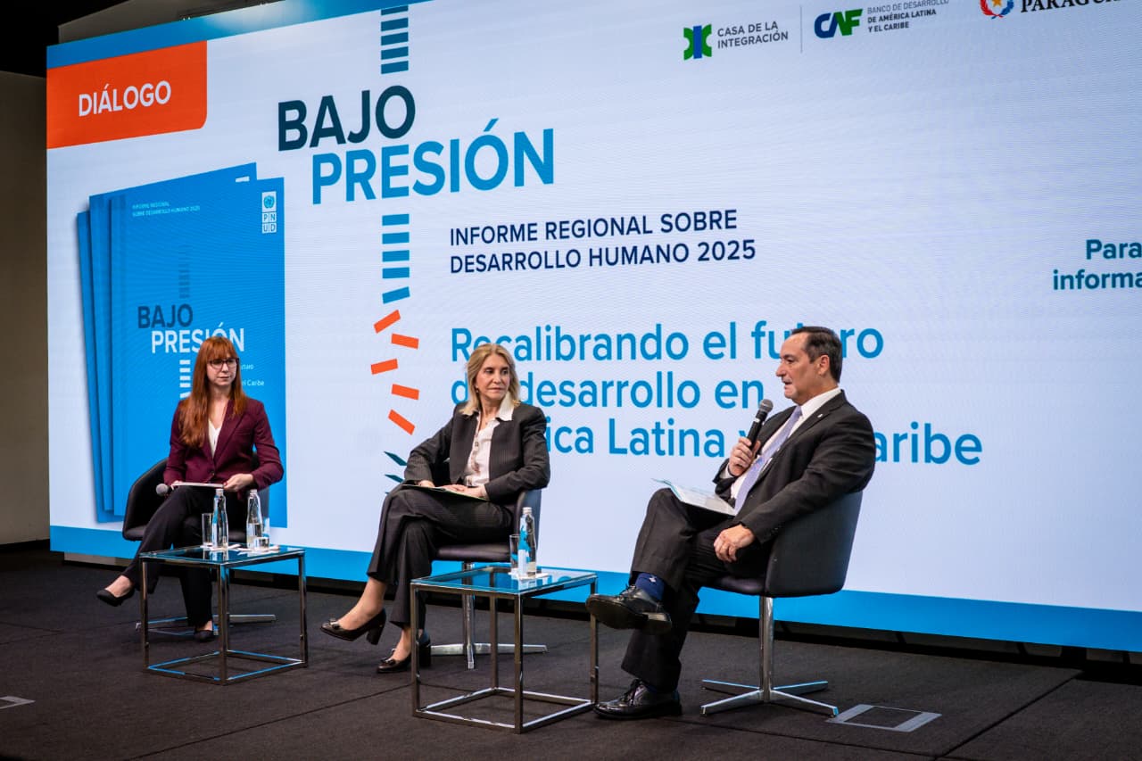 Three panelists seated on stage with microphones, blue backdrop reading 'BAJO PRESIÓN' and logos.